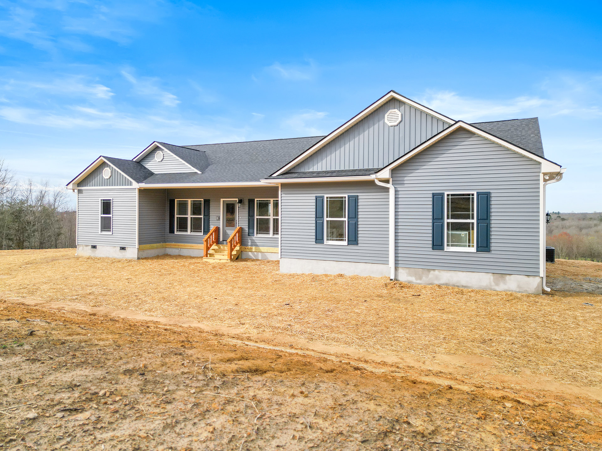 Gravel driveway leading to a cottage-style house with wooden porch stairs, blue-shuttered window, white window frames, and a patch of bare dirt beside grassy yard