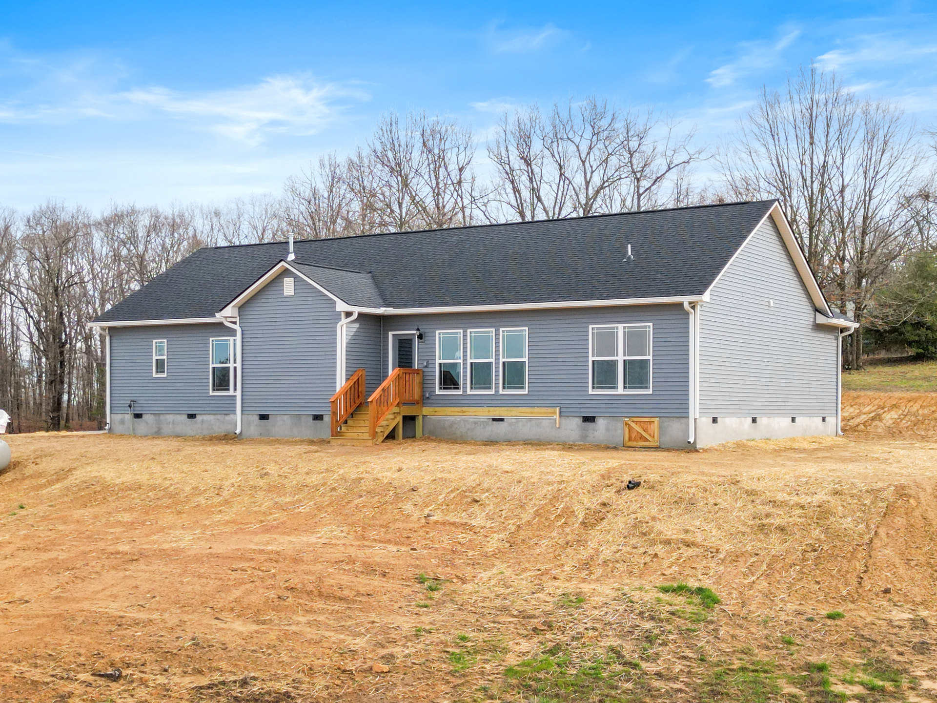 Two-story cottage with white trim windows, wooden exterior staircase, grassy yard, and mature trees under a cloudy sky