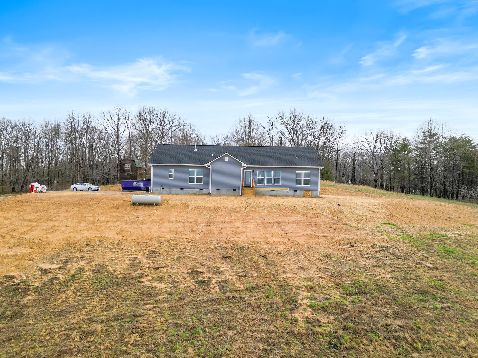 Grey house with white gutters and front stairs, surrounded by a large grassy yard and scattered trees under a blue sky with clouds