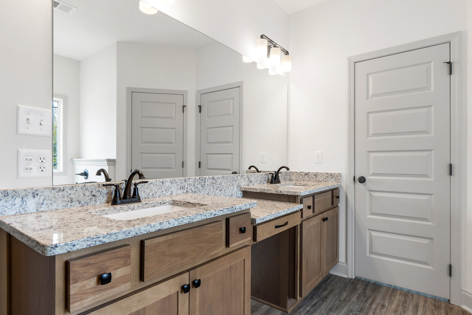 Bathroom with double sinks, black faucets on marble countertops, large mirror above white cabinetry, white door with black hardware, light switch on tiled wall