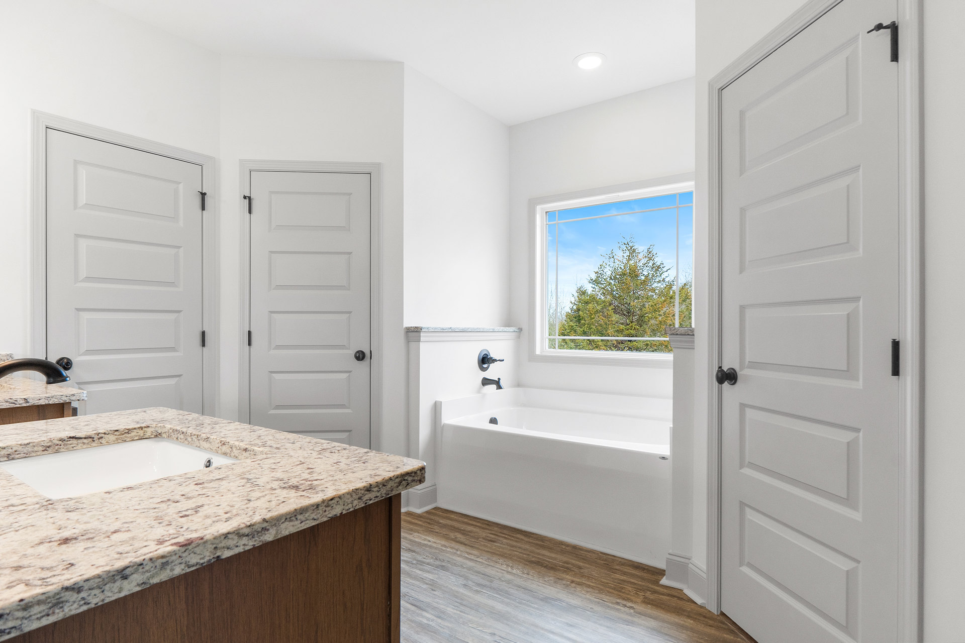 Bathroom with freestanding white bathtub, white countertop sink, light tile flooring, white door with black handle, window overlooking green trees, neutral cabinetry and walls