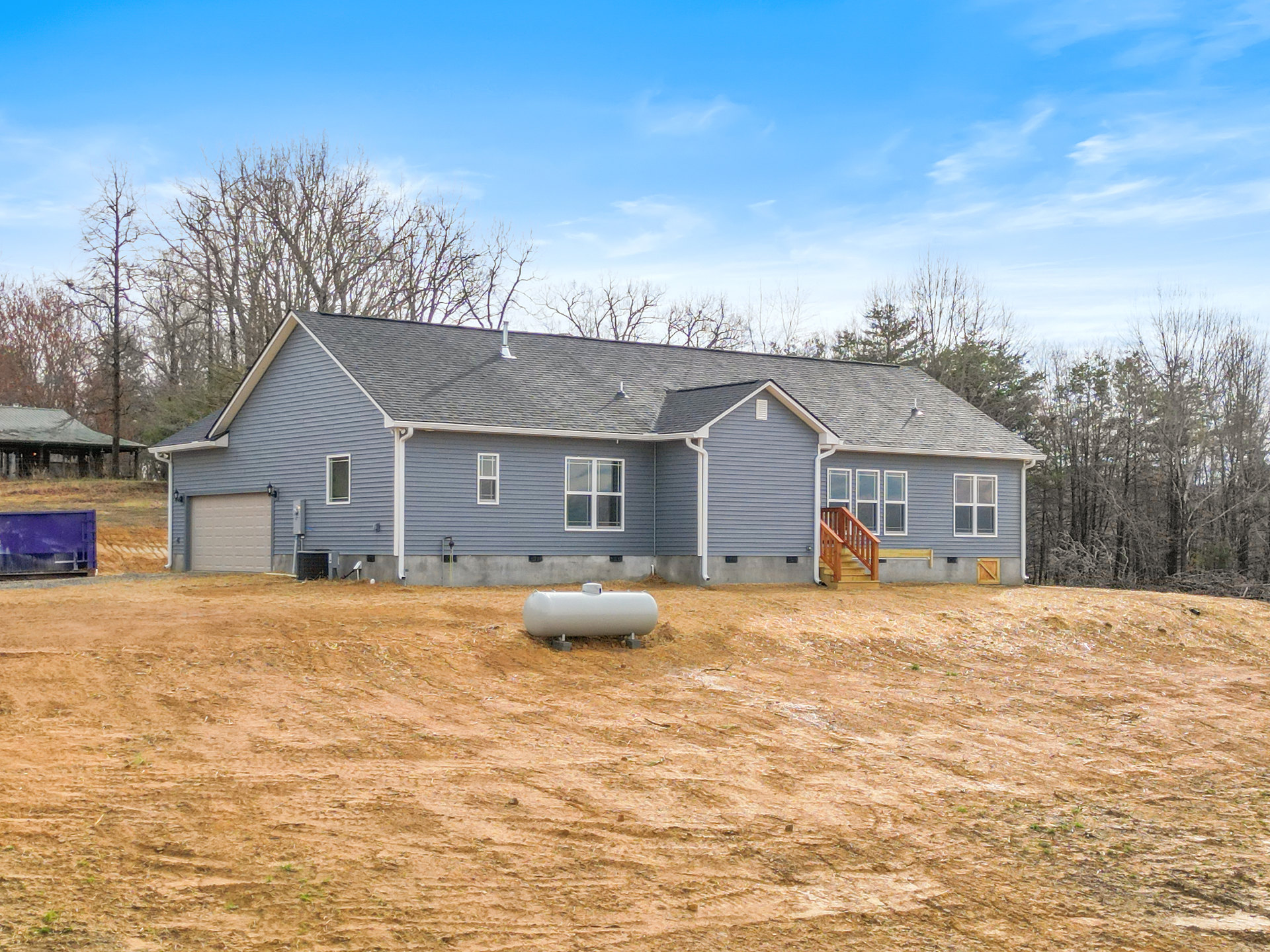 Two-story rural home with gray siding, large paved driveway, wooden staircase railing, white-framed windows, and a white water tank on a dirt lot surrounded by grass and trees.