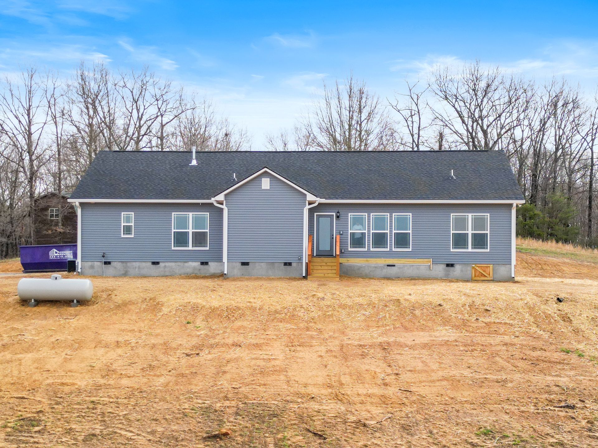 Two-story cottage-style house with covered porch, white-framed windows, front door with stairs, large grassy yard, mature trees, and white gas tank near the entrance