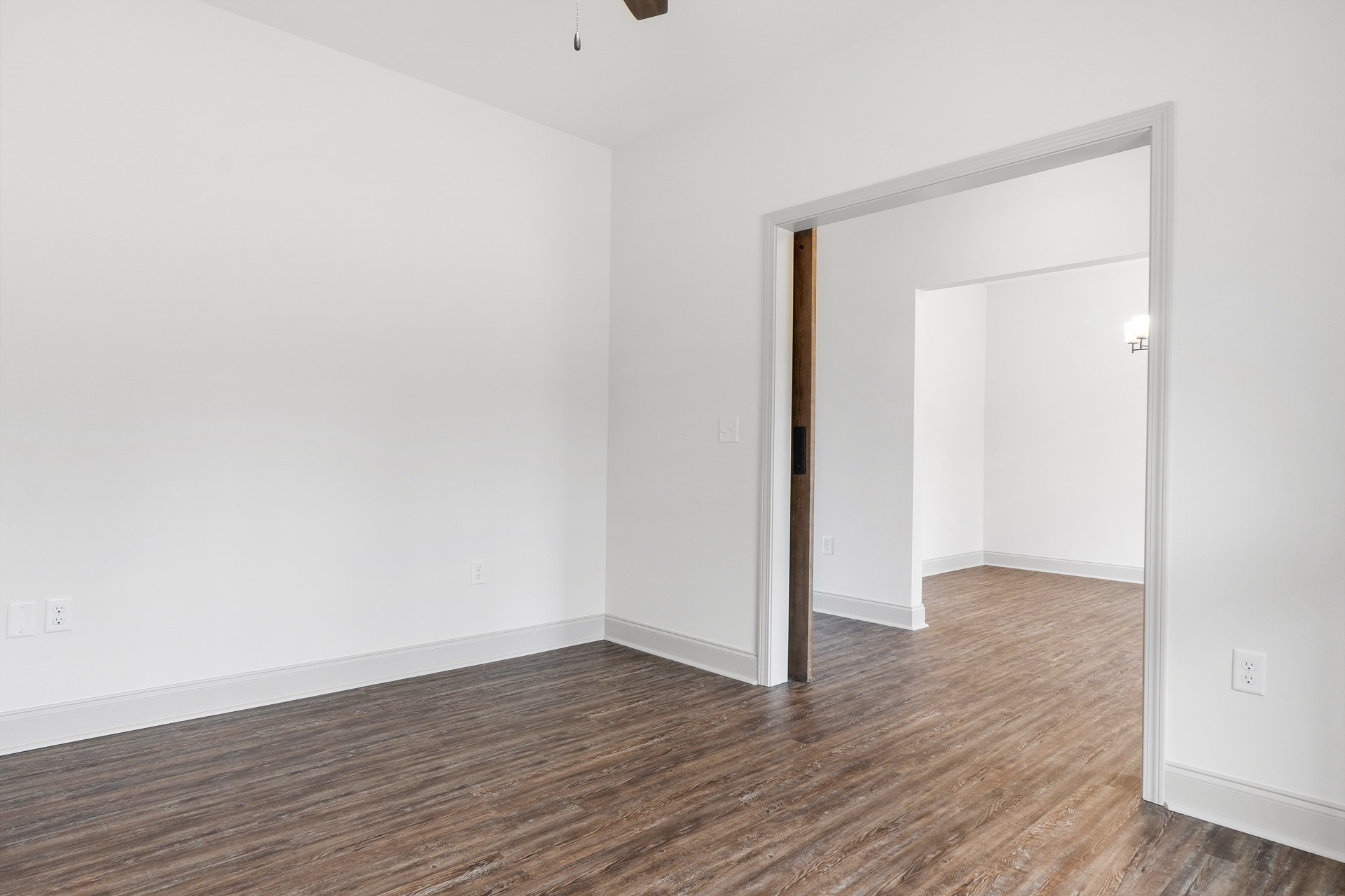 Wood flooring, white walls, white door, ceiling fan, electrical outlet, and light fixture in a bright residential room