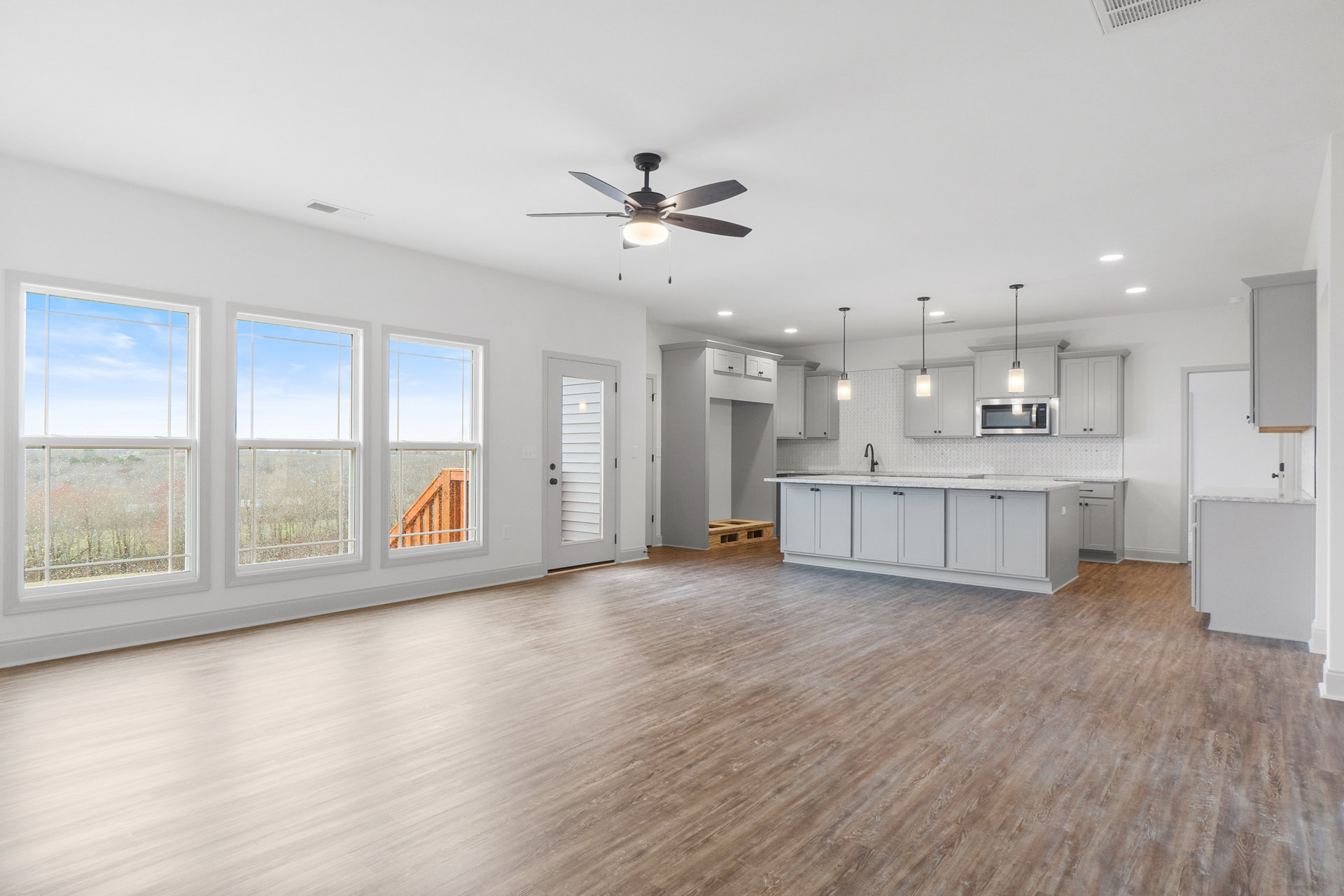 Spacious open room with laminate wood flooring, white walls, ceiling fan with light, adjoining kitchen featuring white cabinets, white countertop with sink, built-in microwave