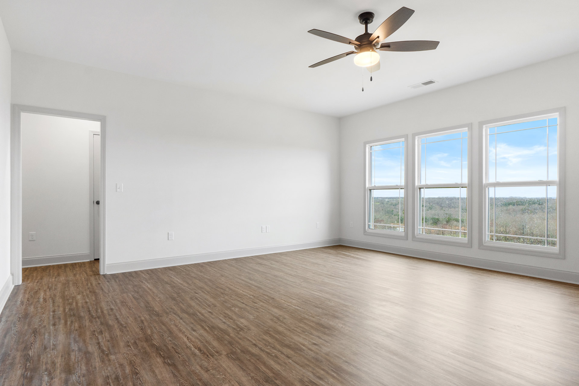 Ceiling fan with light fixture, hardwood floors, row of windows overlooking trees and field, white door with black knob, plaster walls and ceiling