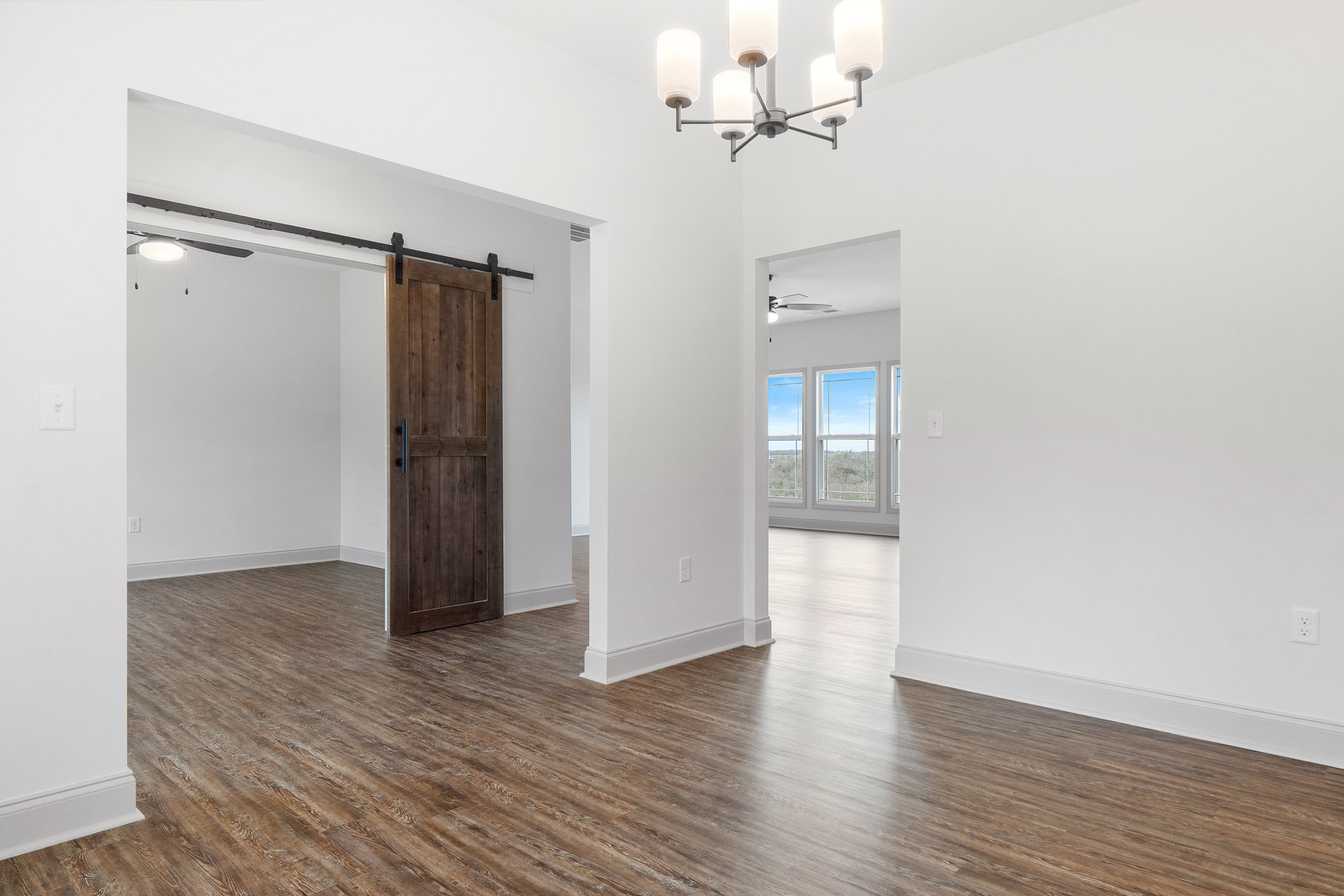 Open room with light wood flooring, white plaster walls, black-handled wooden door, modern chandelier, window showing blue sky, white ceiling fixture, and wall-mounted light switch