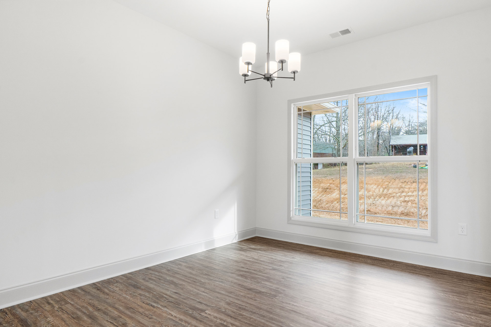 Sunlit room with wide wood plank flooring, large window overlooking farmland, white walls, and modern chandelier hanging from ceiling.