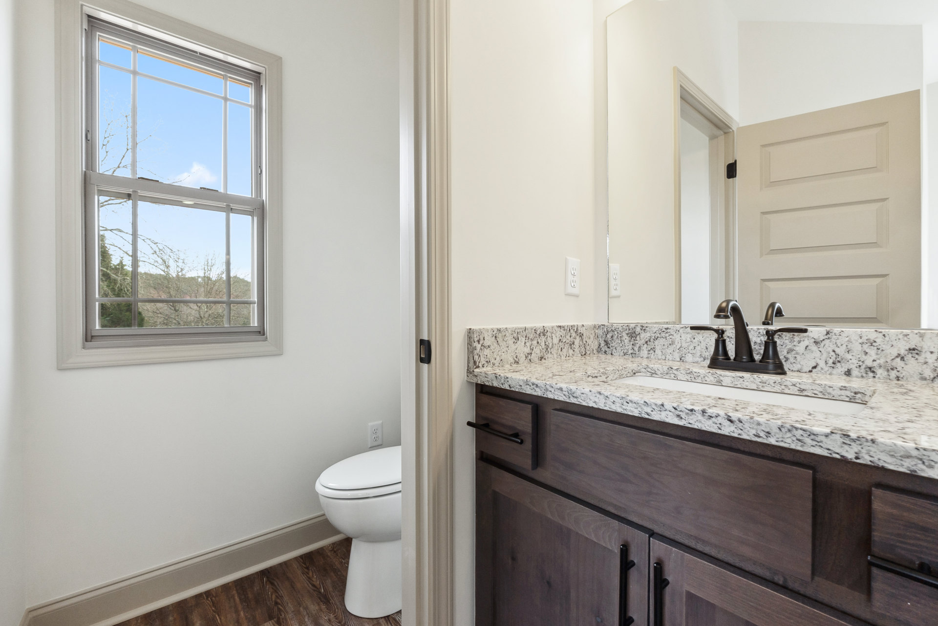 White toilet and rectangular sink with chrome faucet on a light countertop, tile flooring, window showing leafy tree outside, neutral wall color, minimal cabinetry.