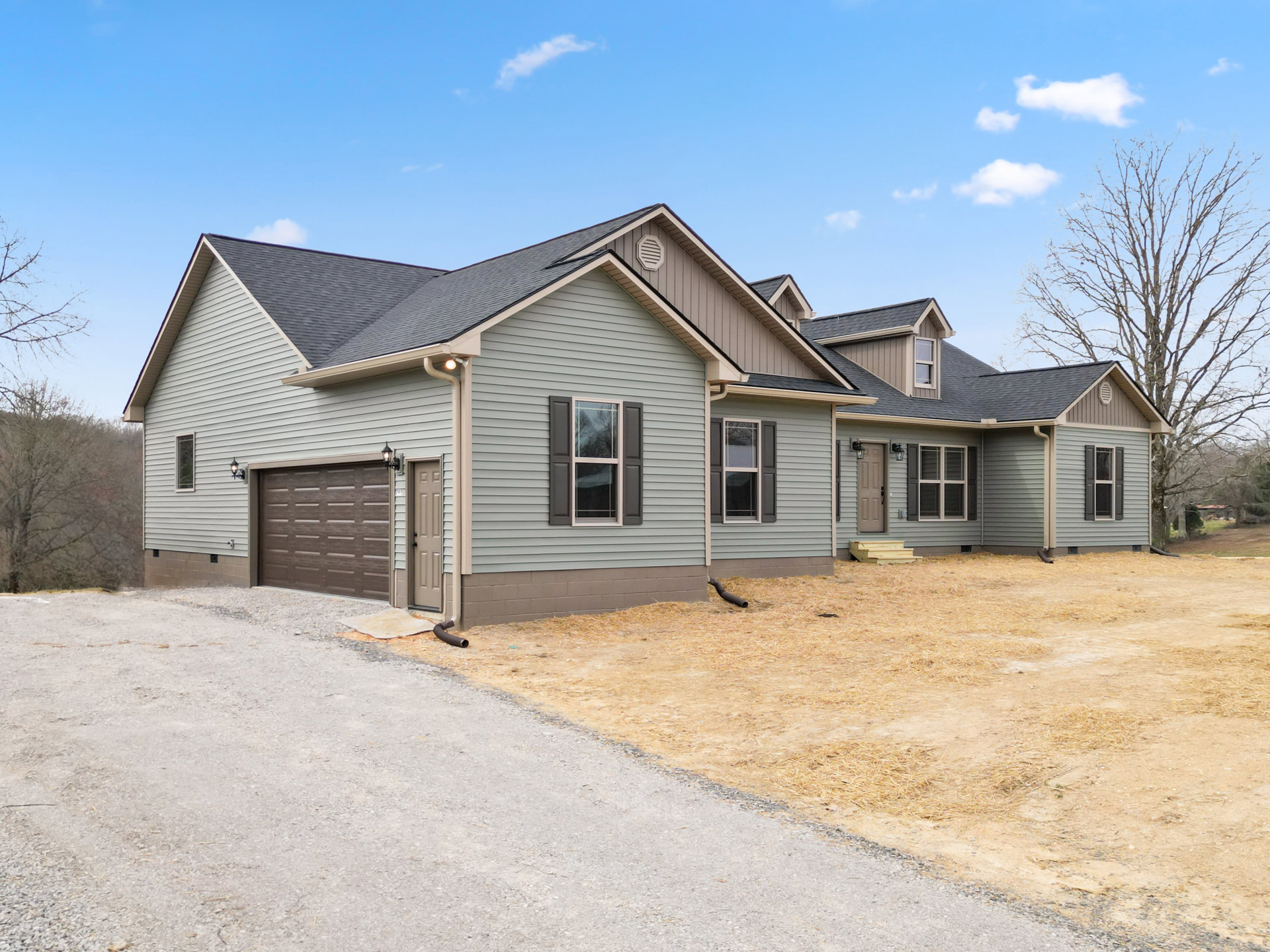 Two-story house with gray siding, white-trimmed windows reflecting trees, attached garage with side light, paved driveway, and front yard landscaping.