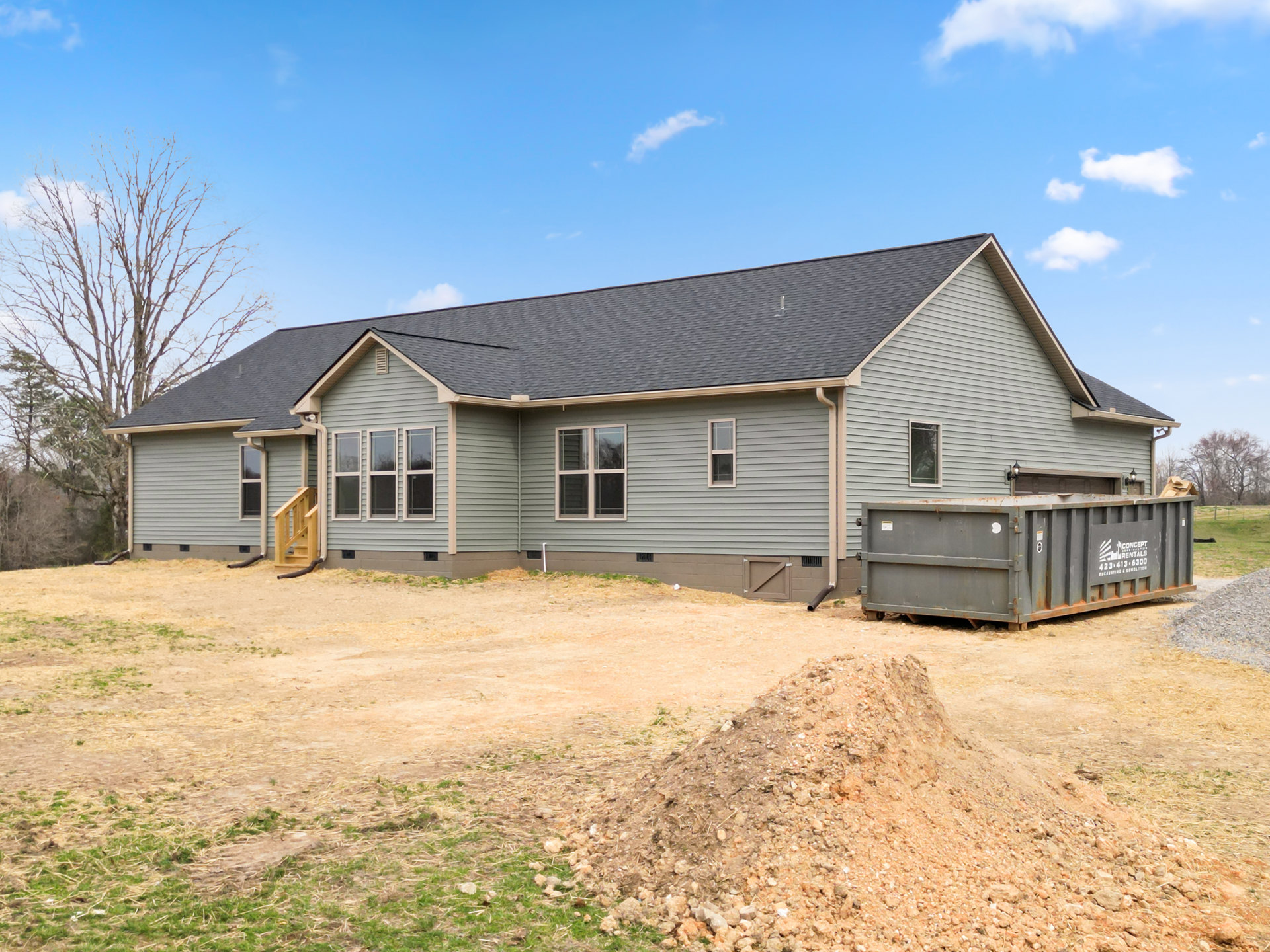 Two dirt piles beside a house with a grey roof, white-framed window, and wooden staircase railing; rusted grey container visible outdoors under cloudy sky.
