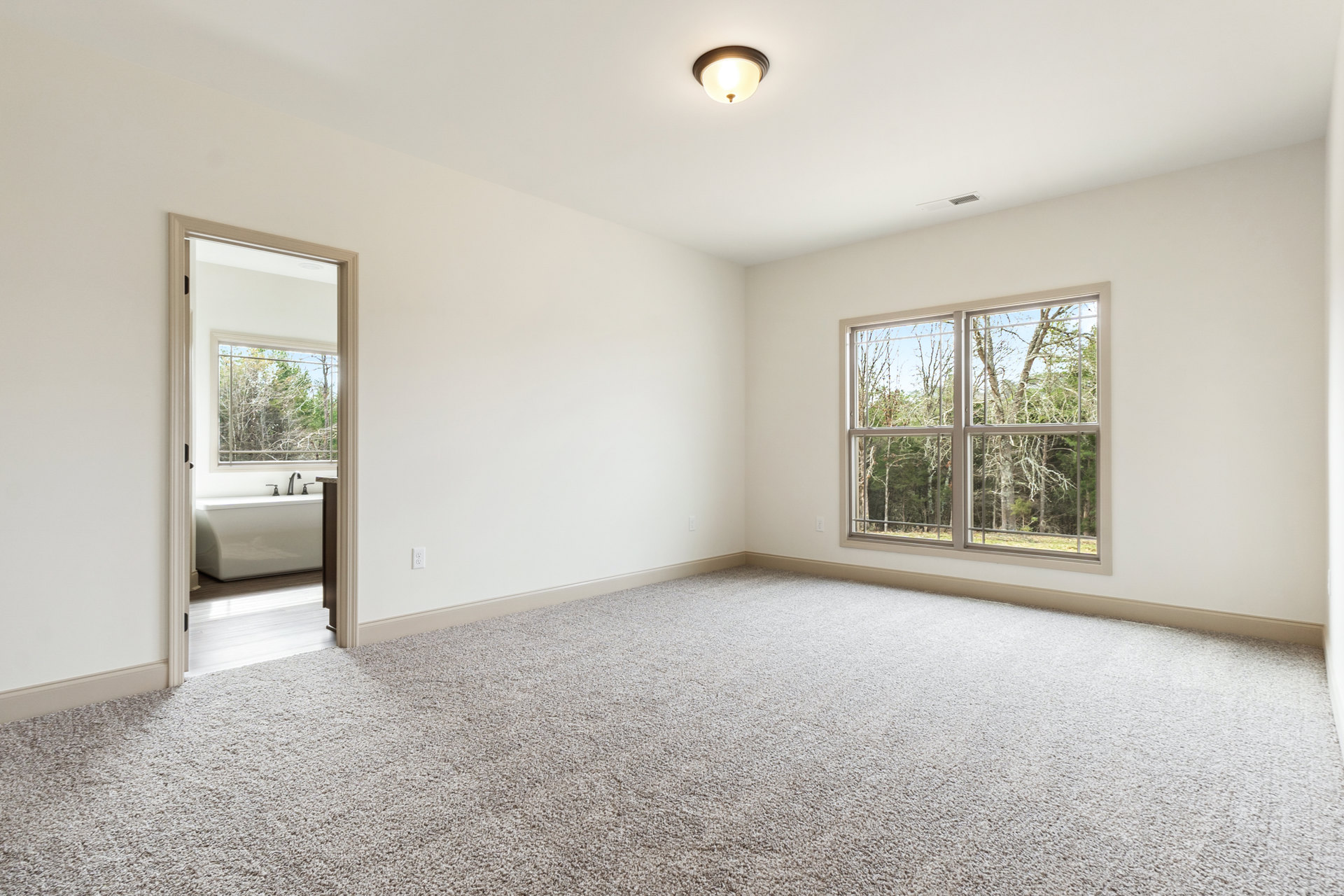 Bathroom with white freestanding tub, chrome faucets, large window overlooking trees, light fixture, white walls, and carpeted floor