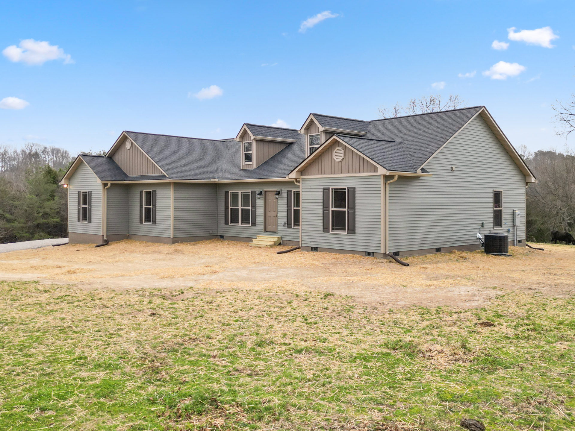 Two-story wood-sided house with white-framed windows, grassy yard, mature trees, and blue sky with scattered clouds