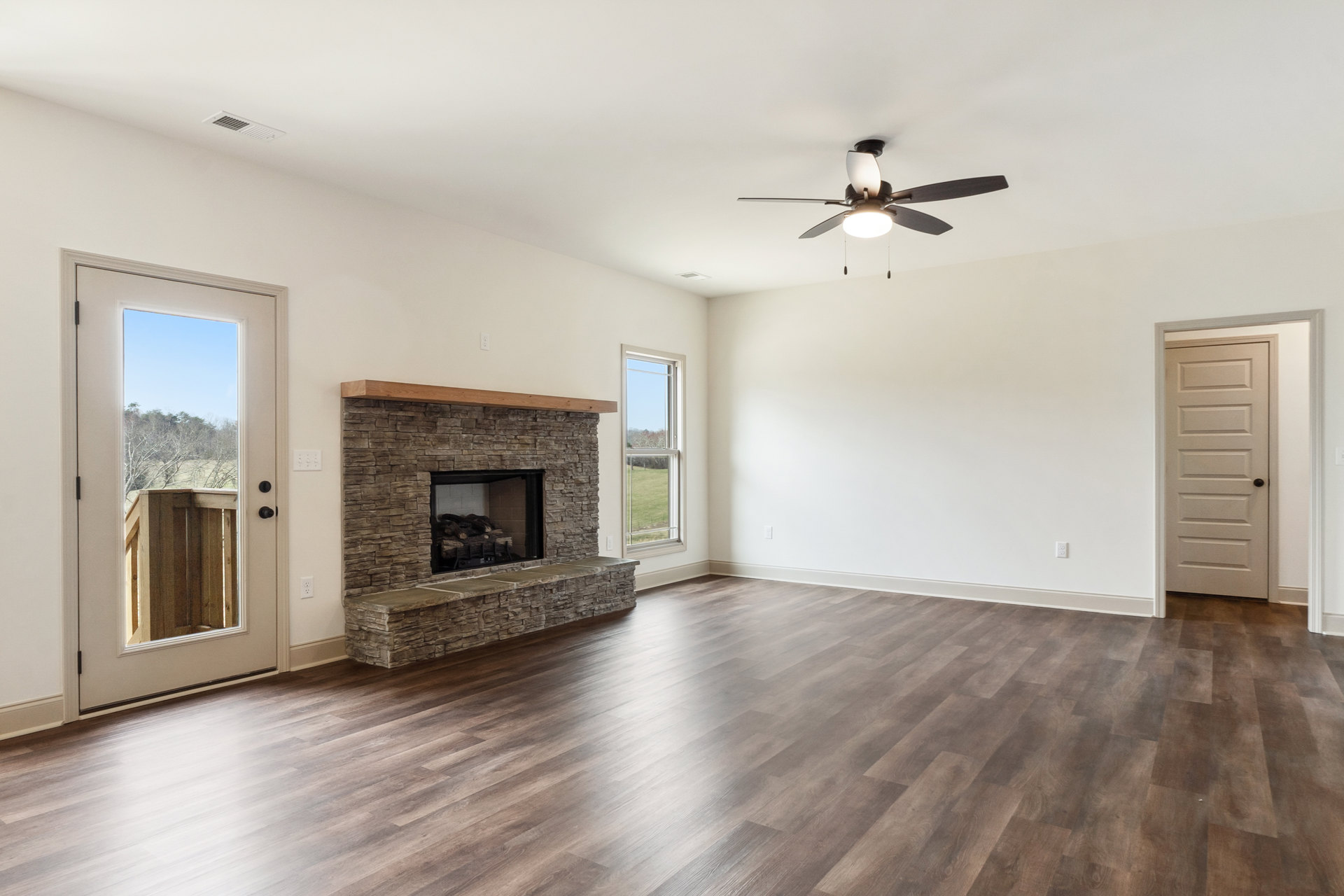 Living room with wood flooring, stone fireplace burning wood, ceiling fan with light, large window and door offering views of trees and blue sky.