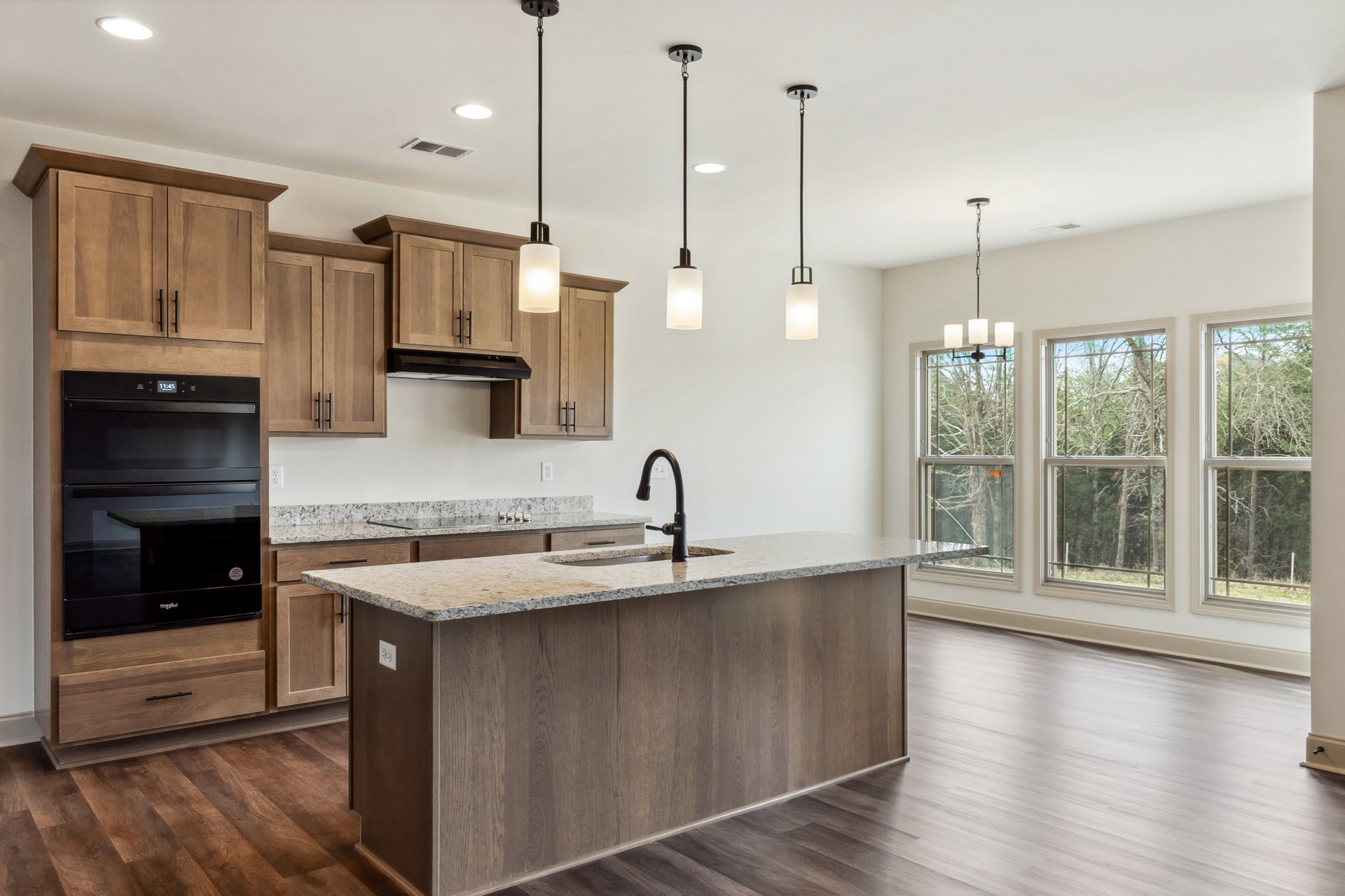 Kitchen with wood floors, white cabinetry, granite countertops, central island with built-in sink, black glass-door oven, pendant lighting, large windows overlooking trees