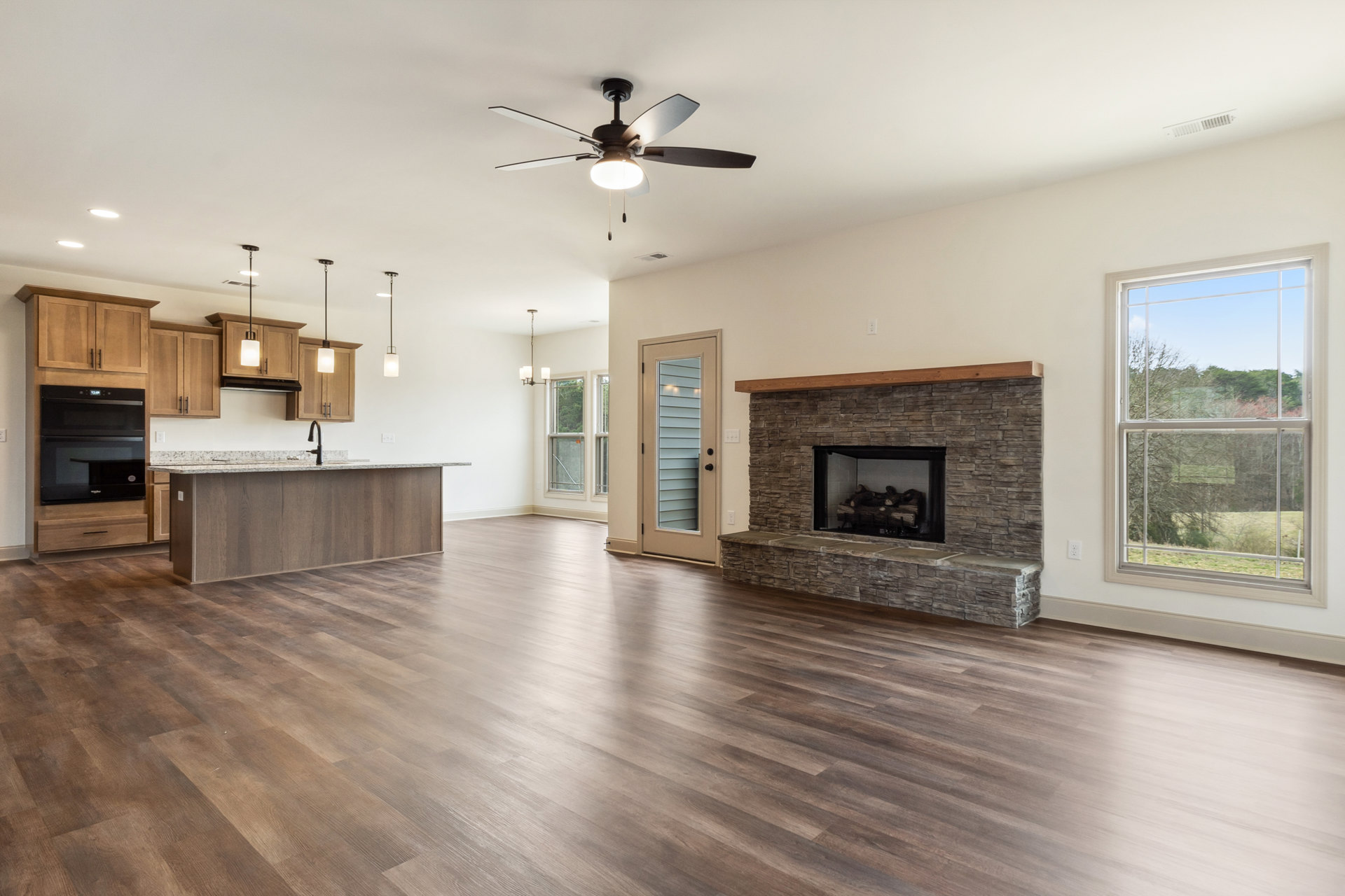 Living room with wood flooring, central fireplace burning logs, ceiling fan with light fixture, large window overlooking trees, black microwave oven mounted on wall