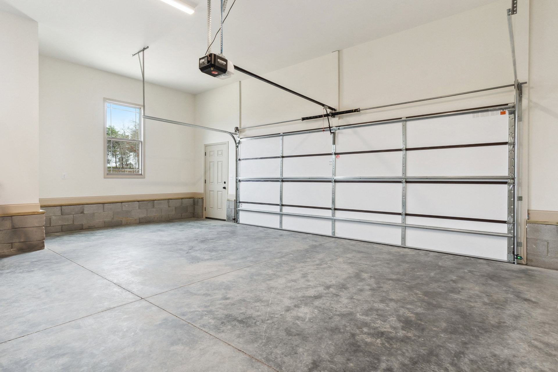 Garage interior with smooth concrete floor, white plaster walls, overhead garage door, side window showing green trees, white entry door with black hardware, metal shelving, and a