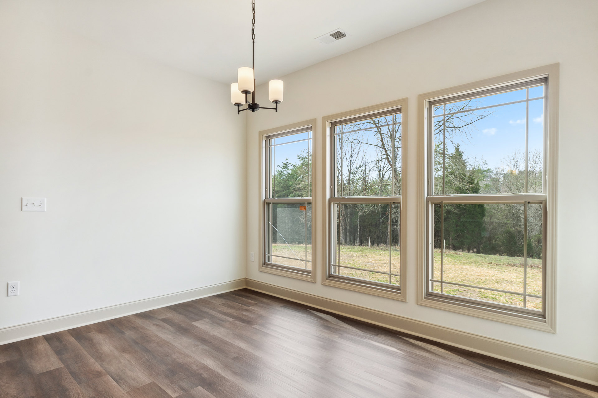 Chandelier hanging from ceiling above wood flooring, large windows with views of trees, white walls, daylight illuminating interior