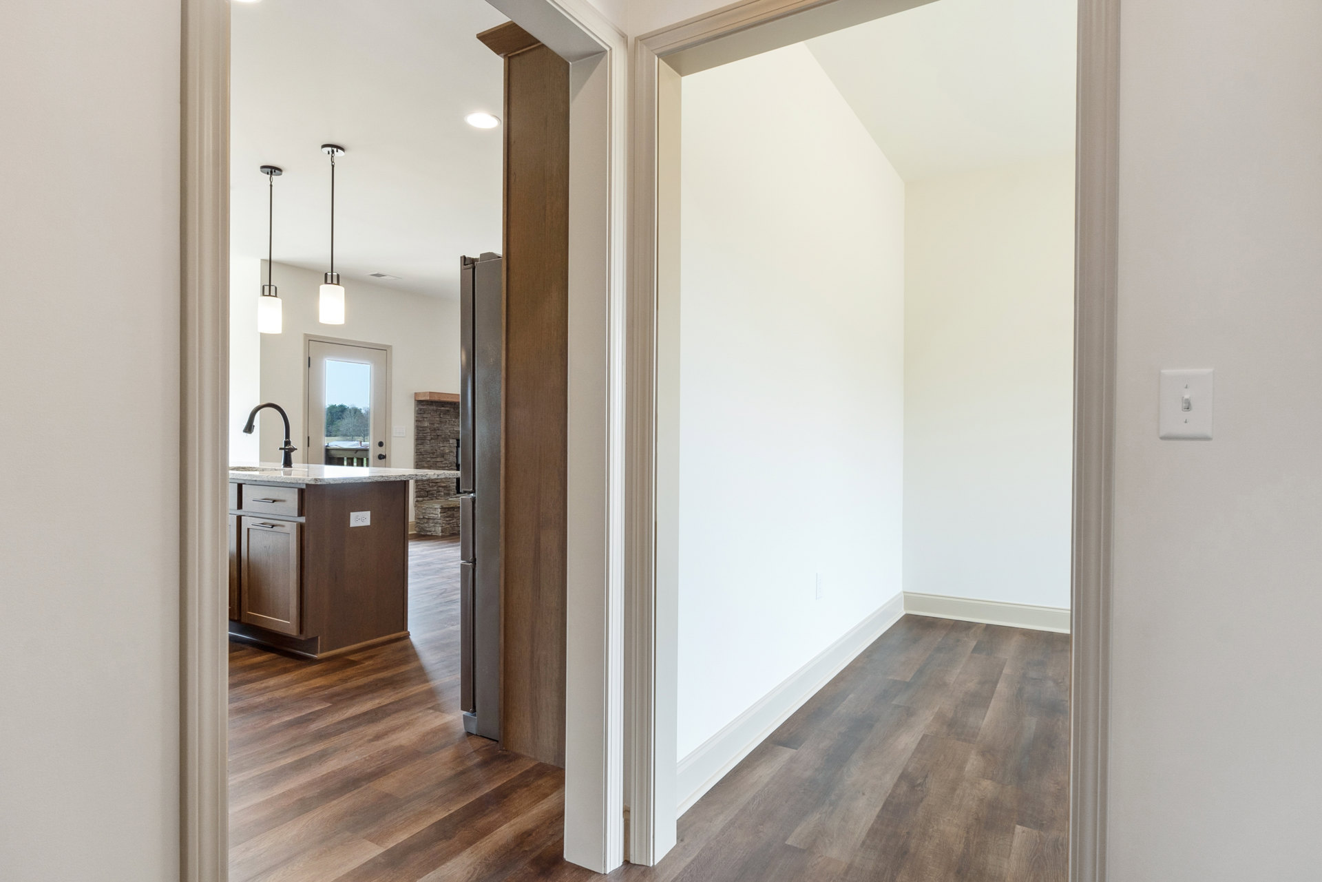 Hallway with wood flooring leading to a kitchen featuring a marble-topped island, white walls, refrigerator, light switch, and a white door with a window