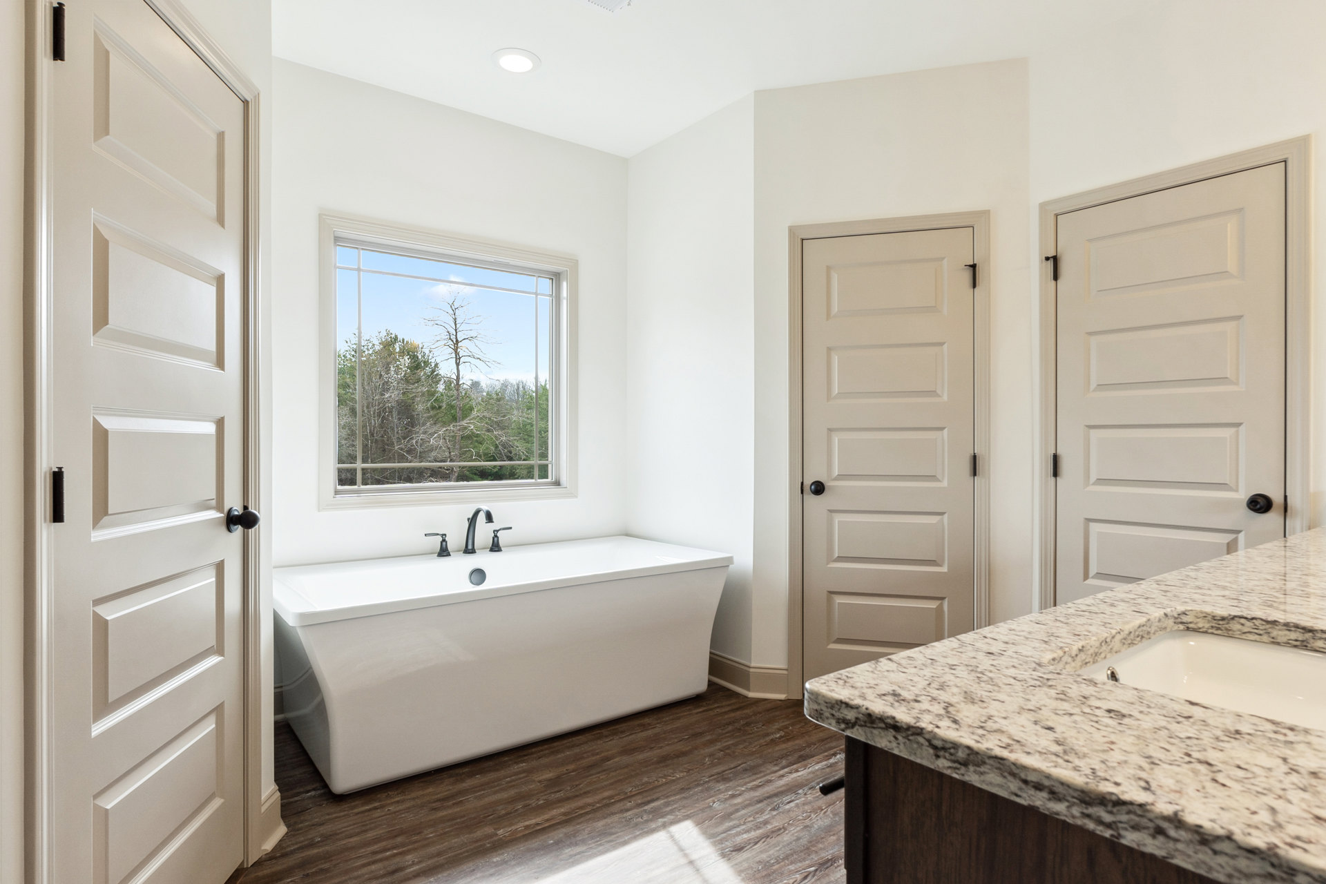 White bathtub with black faucets beside a marble countertop and white sink, window showing trees outside, white door with black knob, light tile flooring and walls