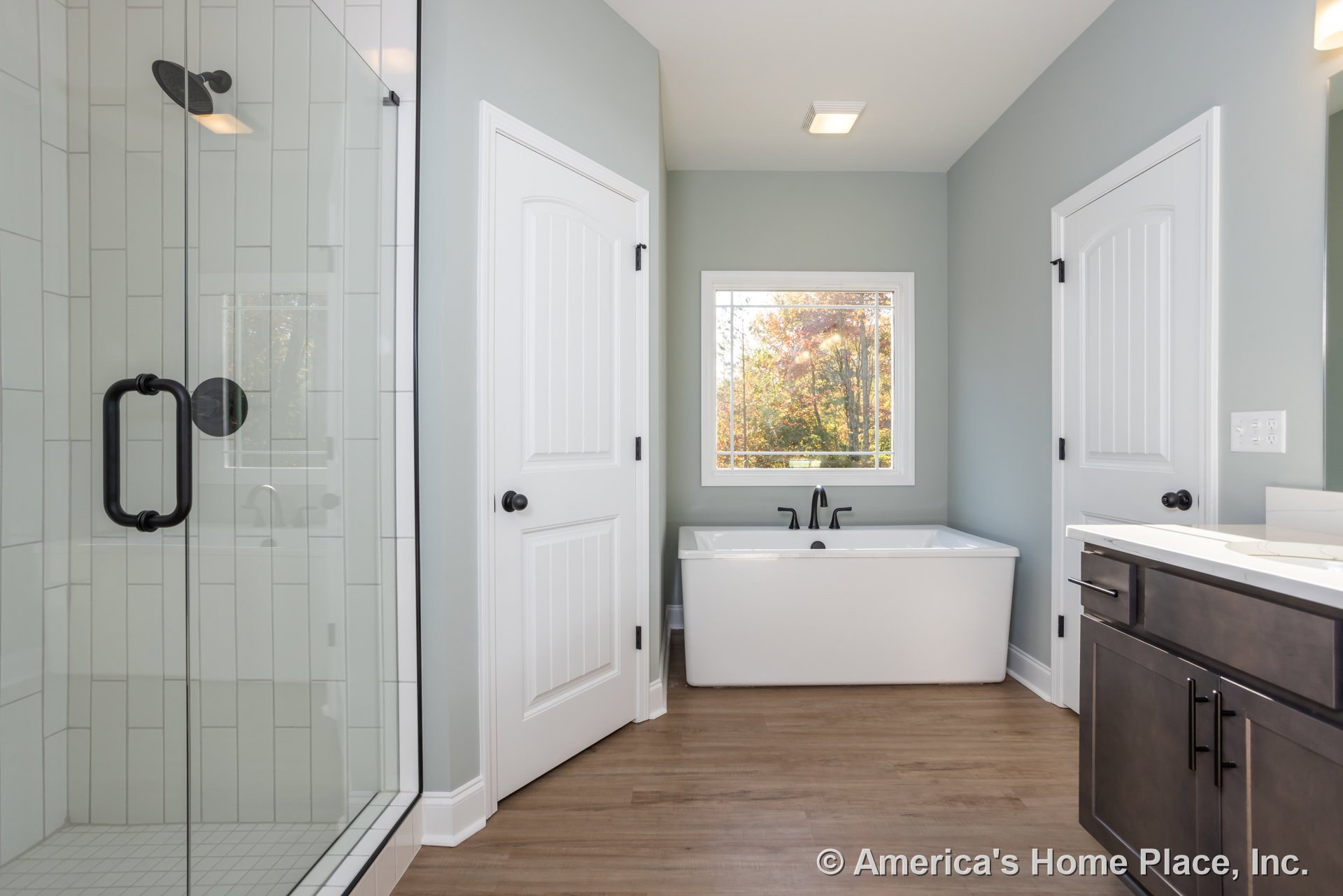 White bathtub with matte black faucet and handle, white tile shower wall, window overlooking trees, close-up of white cabinet and electrical outlet with switches