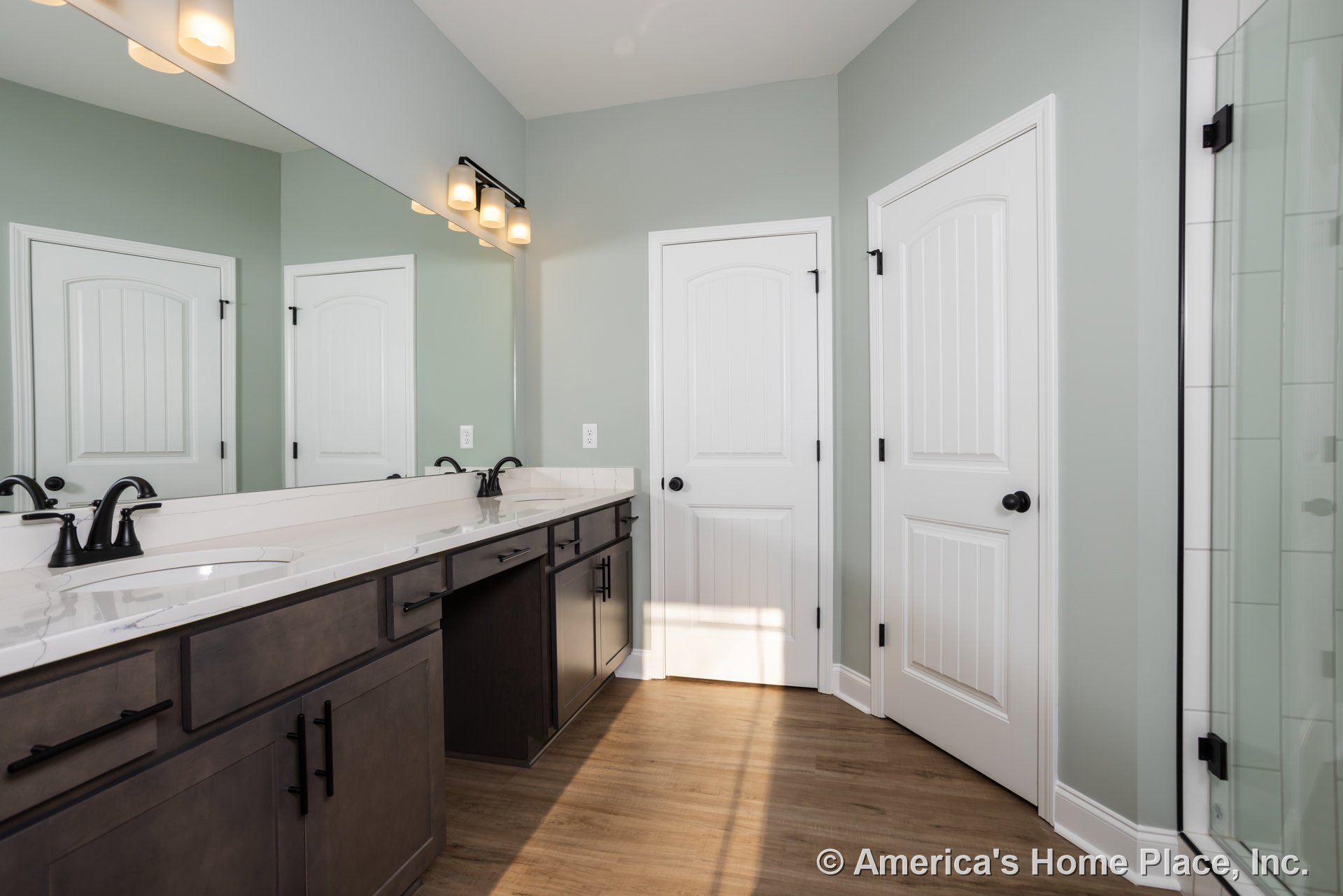 Bathroom with double vanity, rectangular mirror above sinks, white door with black hardware, tile flooring, and partial view of glass shower door