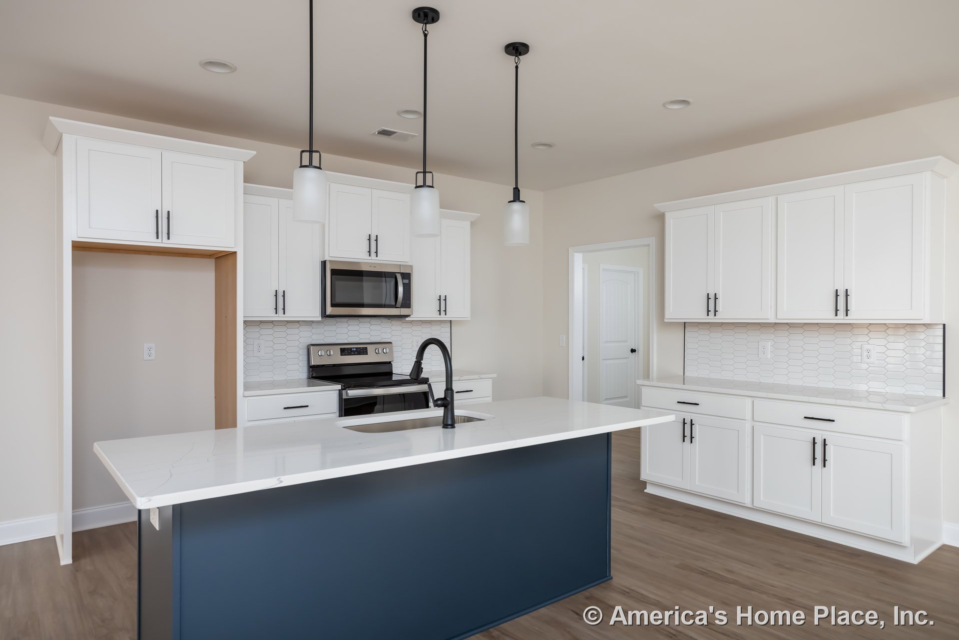 White shaker cabinets and blue island with quartz countertop, open microwave door, black pendant lamp, stainless steel appliances, white backsplash, and decorative bottle on