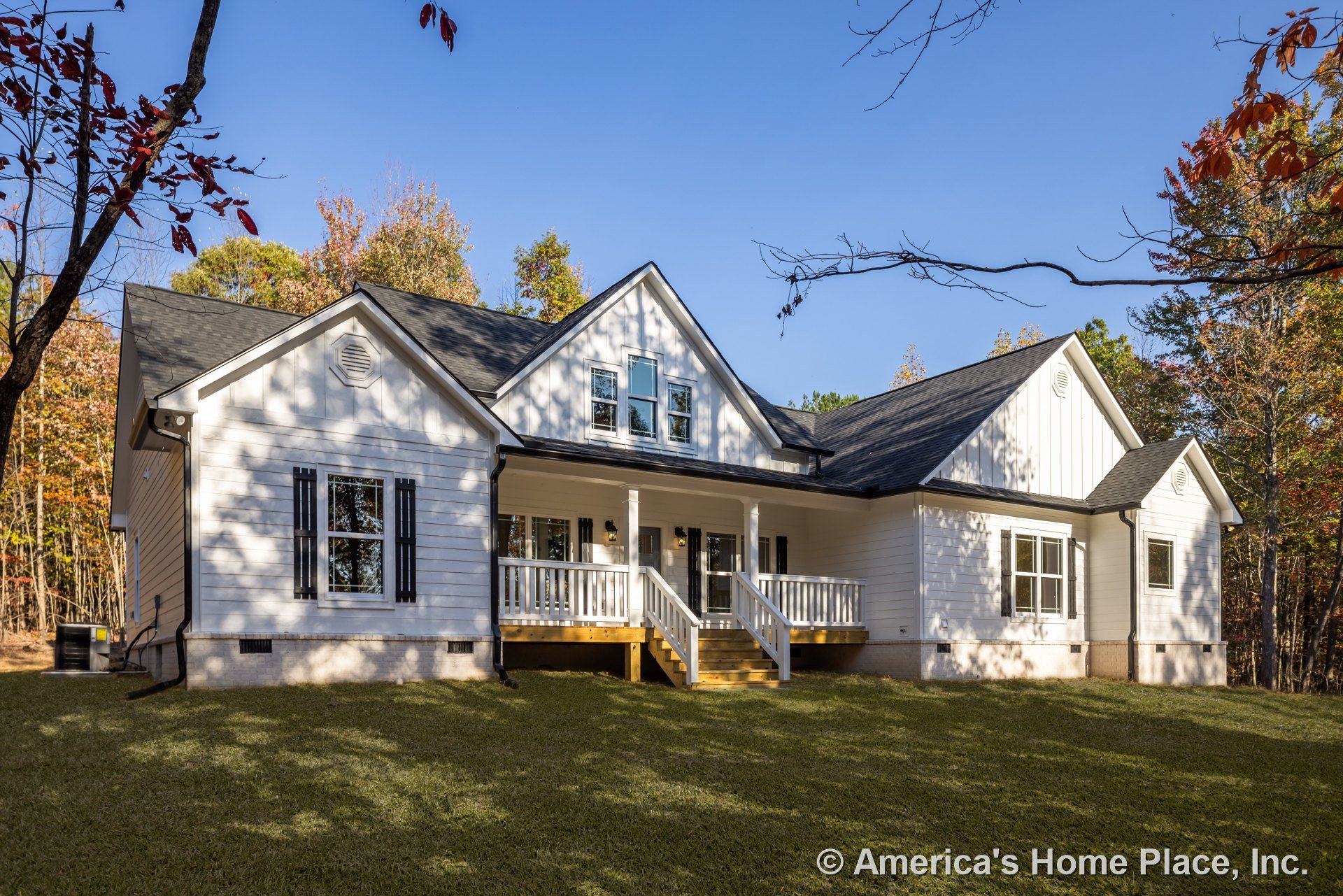 White two-story house with black shutters, front porch, wooden stairs, green lawn, and mature trees in the background
