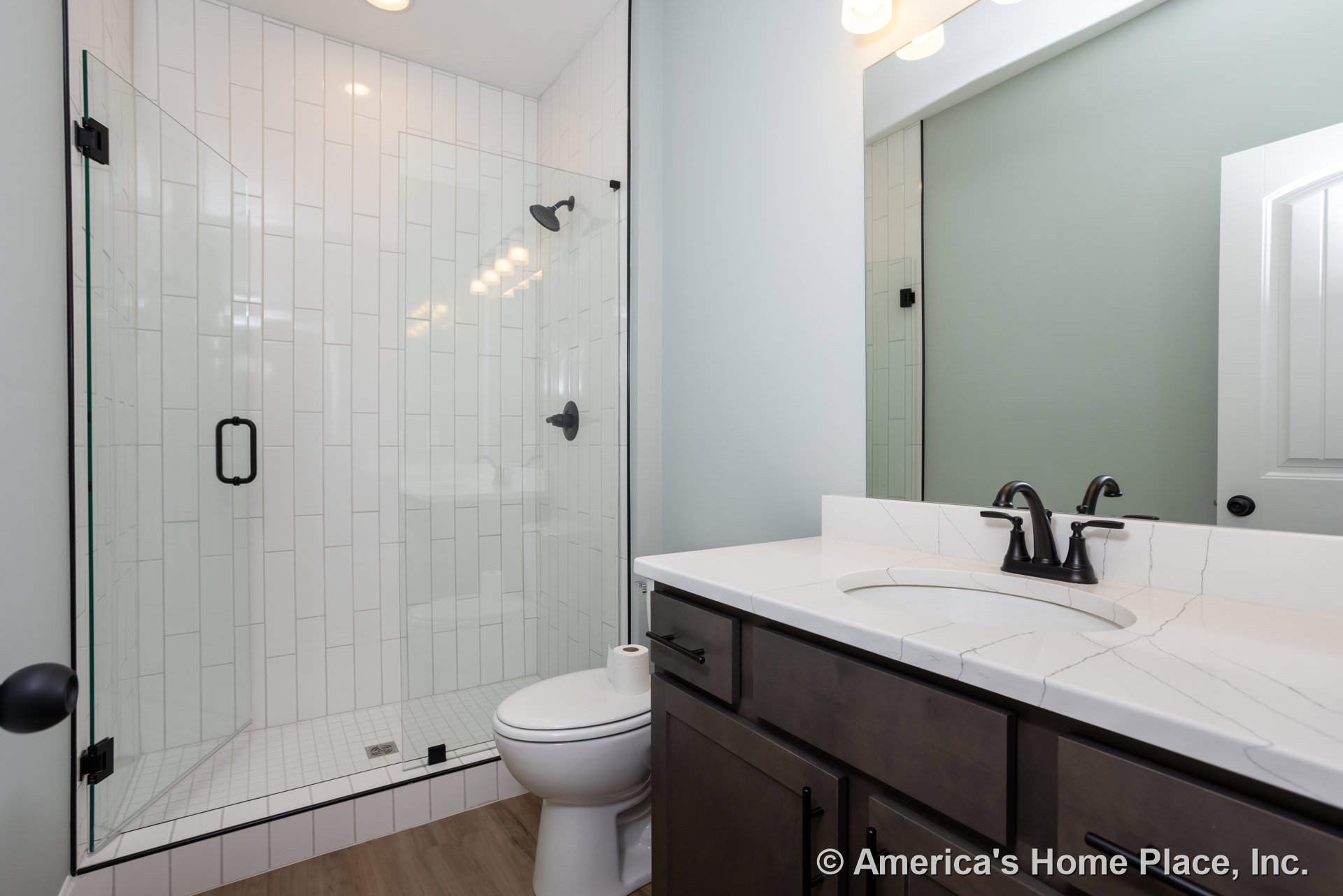 Modern bathroom with white tile walls, glass-enclosed shower, chrome shower head, white vanity with integrated sink, toilet with toilet paper roll on lid, black metal towel holder