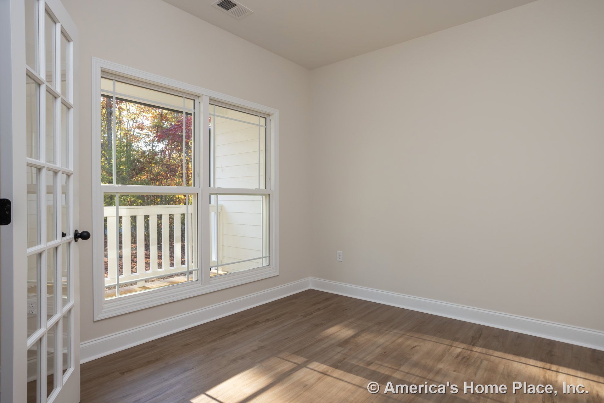 Wood flooring with white baseboard trim, large window overlooking leafy trees, white railing visible outside.