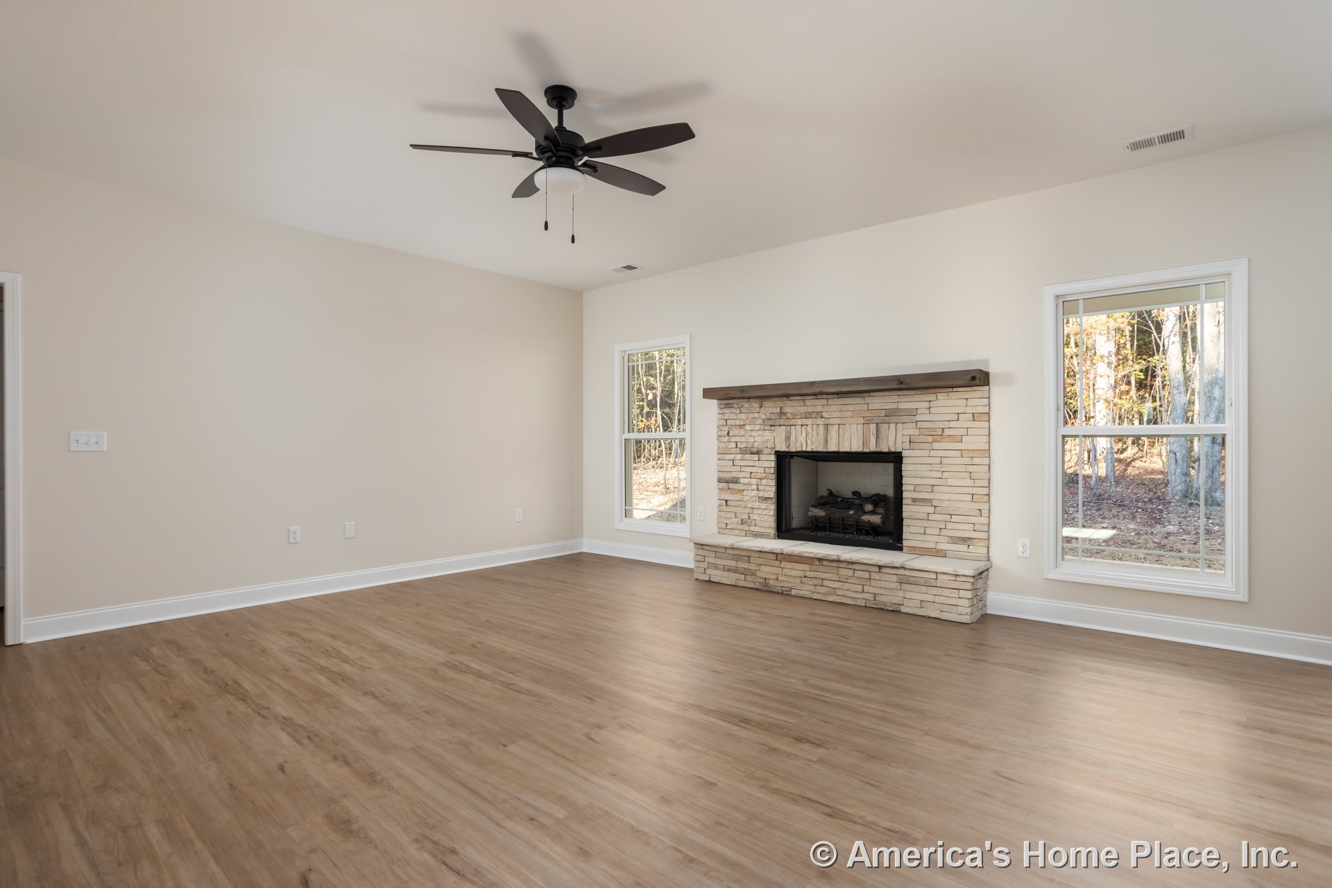 Living room with wood flooring, central fireplace with burning log, ceiling fan with light fixture, large window overlooking trees.