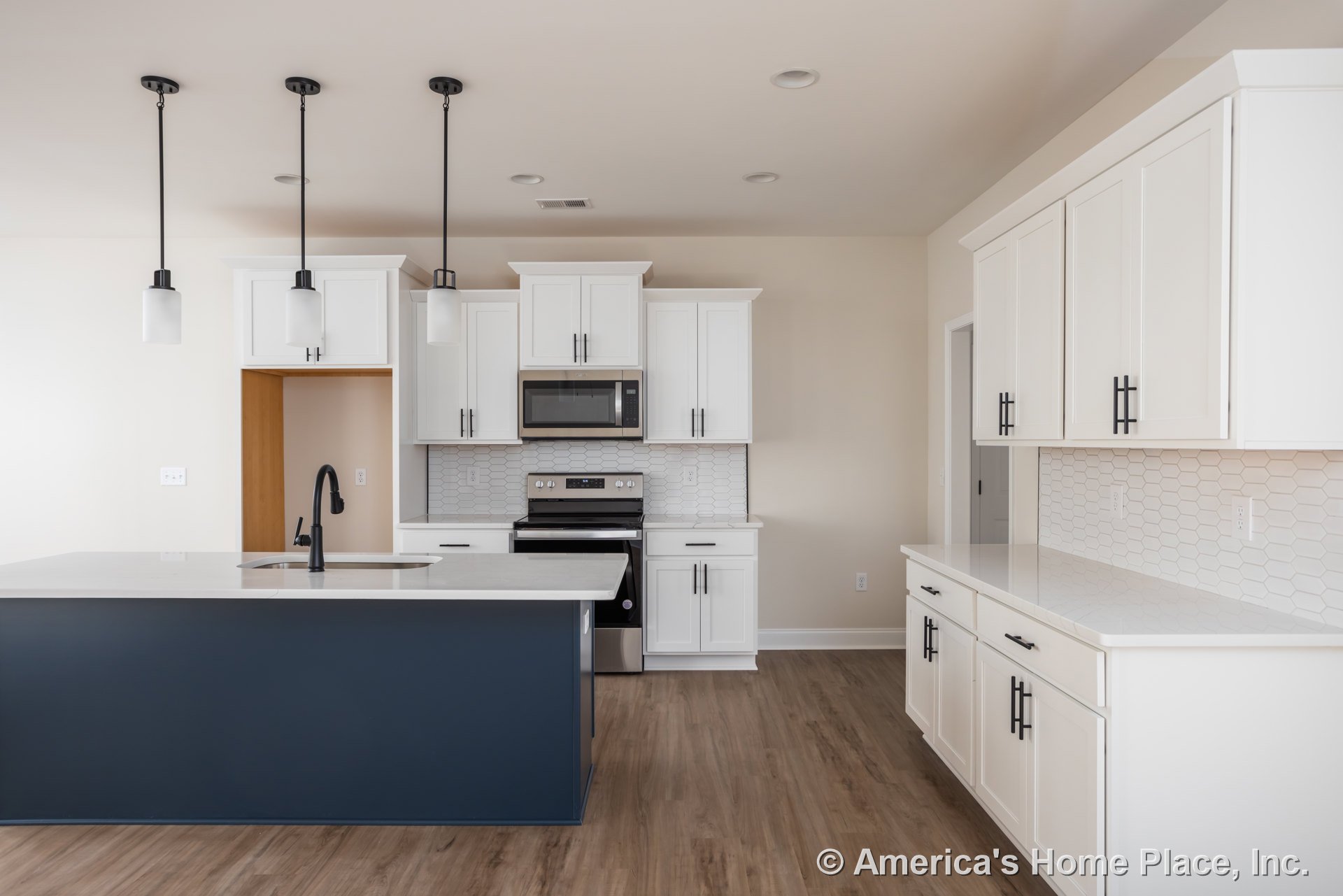 White kitchen cabinets with black handles, blue island with sink, open microwave oven, black and silver stove, light countertops, wood flooring, white walls.