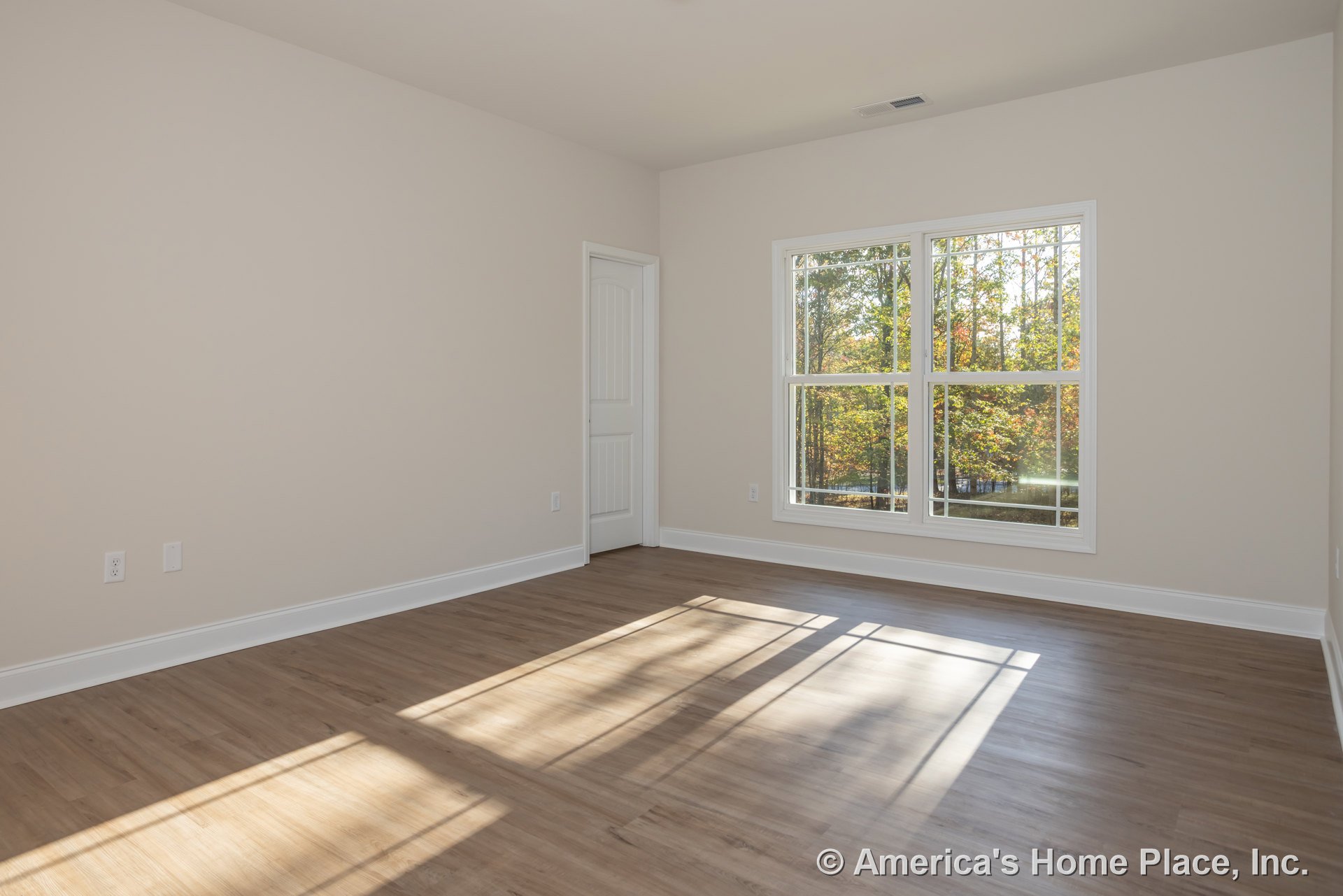 Wood flooring in a bright room with a large window overlooking trees, white walls, and a white door with a black handle