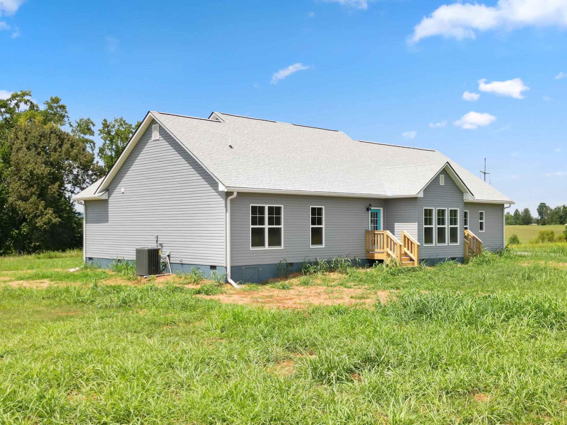Two-story farmhouse with white siding, covered front porch, wooden exterior stairs, white-framed windows, fenced yard, green grass, mature trees, and cloudy sky in the background