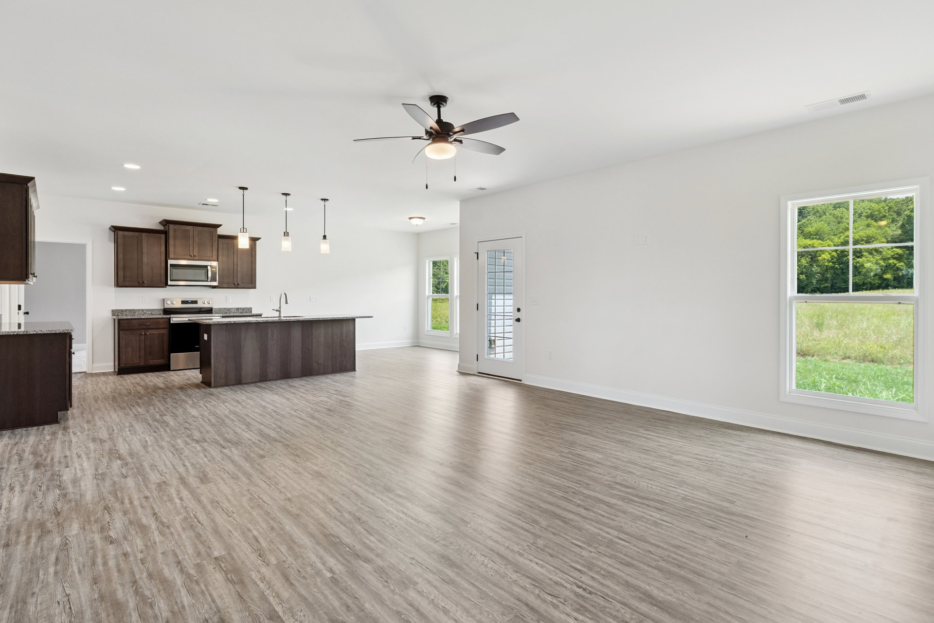 Spacious kitchen and living area with wood flooring, central kitchen island, ceiling fan with light, open microwave, white cabinetry, glass-panel door, and window overlooking