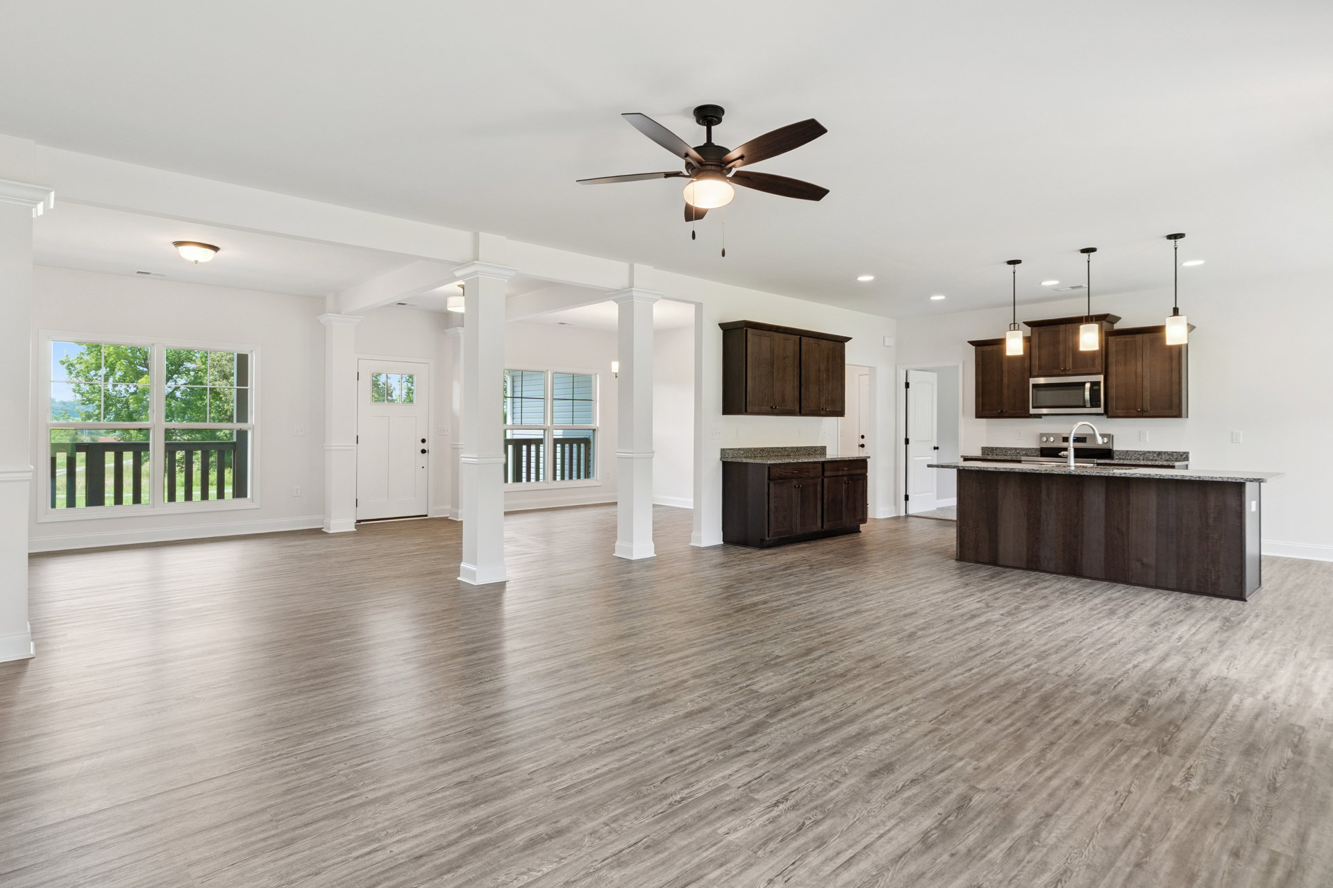 Spacious open floor plan featuring wood flooring, white pillars, kitchen island with silver appliance, ceiling fan with light, open microwave door, and window overlooking trees.
