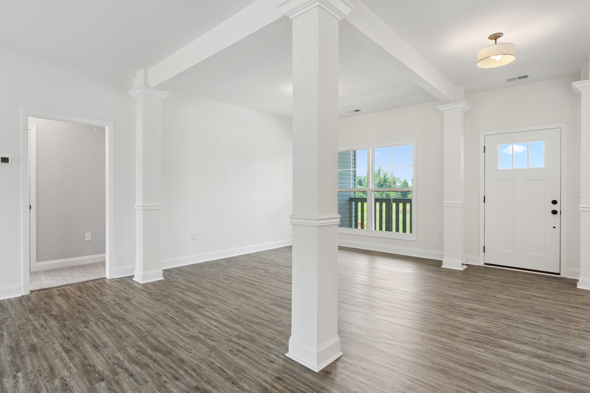 Hardwood floor room with white pillars, white door with window, white column, light fixture with white shade, window overlooking fenced grassy yard
