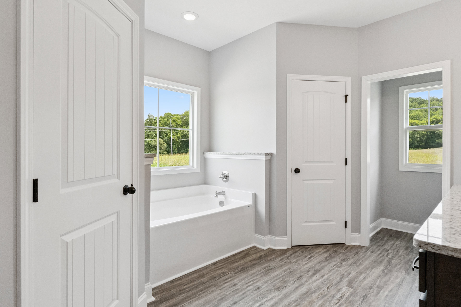 Bathroom with white walls, freestanding white bathtub with silver faucet, wood floor with white trim, window showing trees, white door with black handle