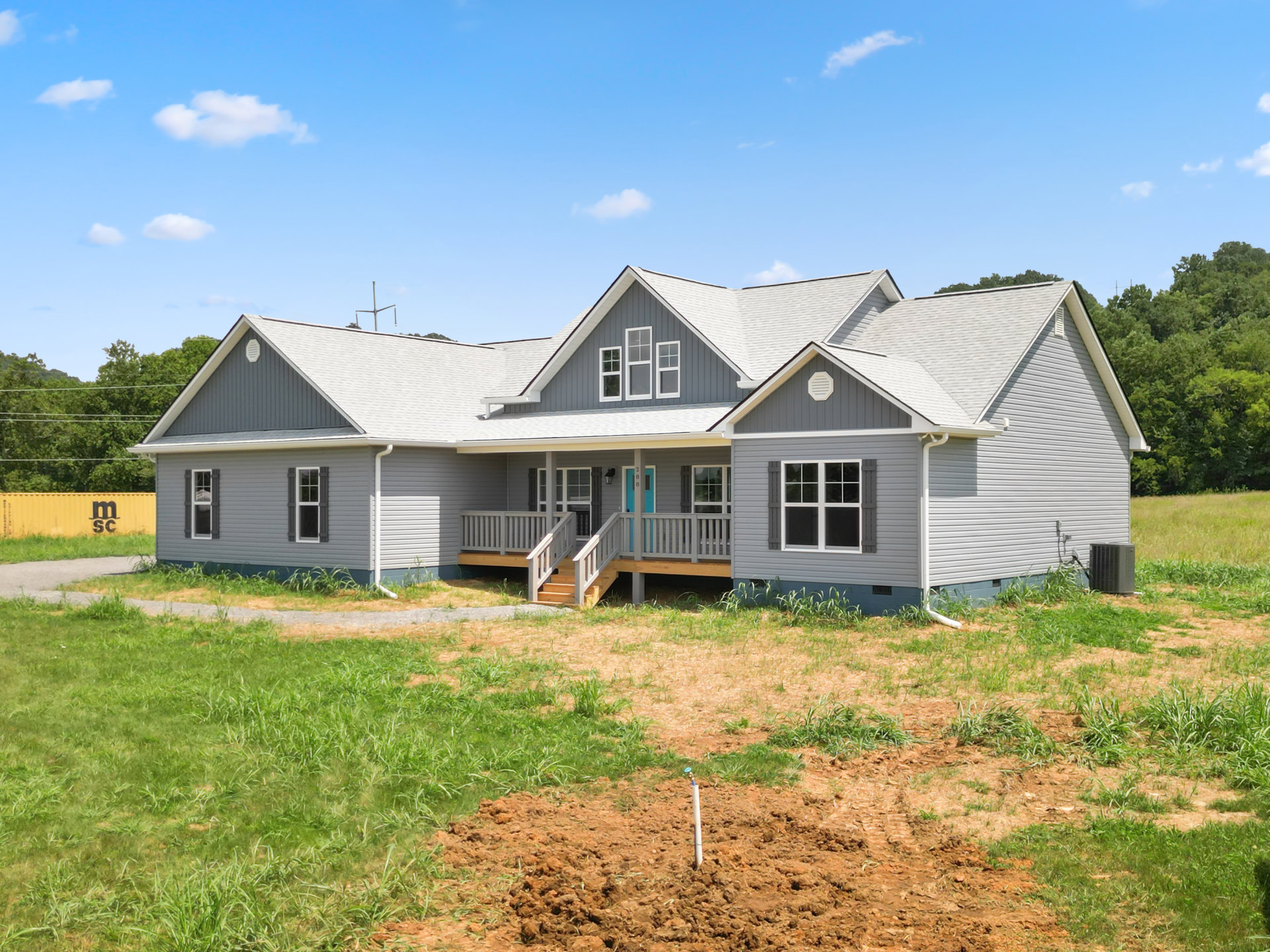 Two-story house with blue front door, white siding, window shutters, wooden steps, green lawn, grass field, and sprinkler under a clear blue sky