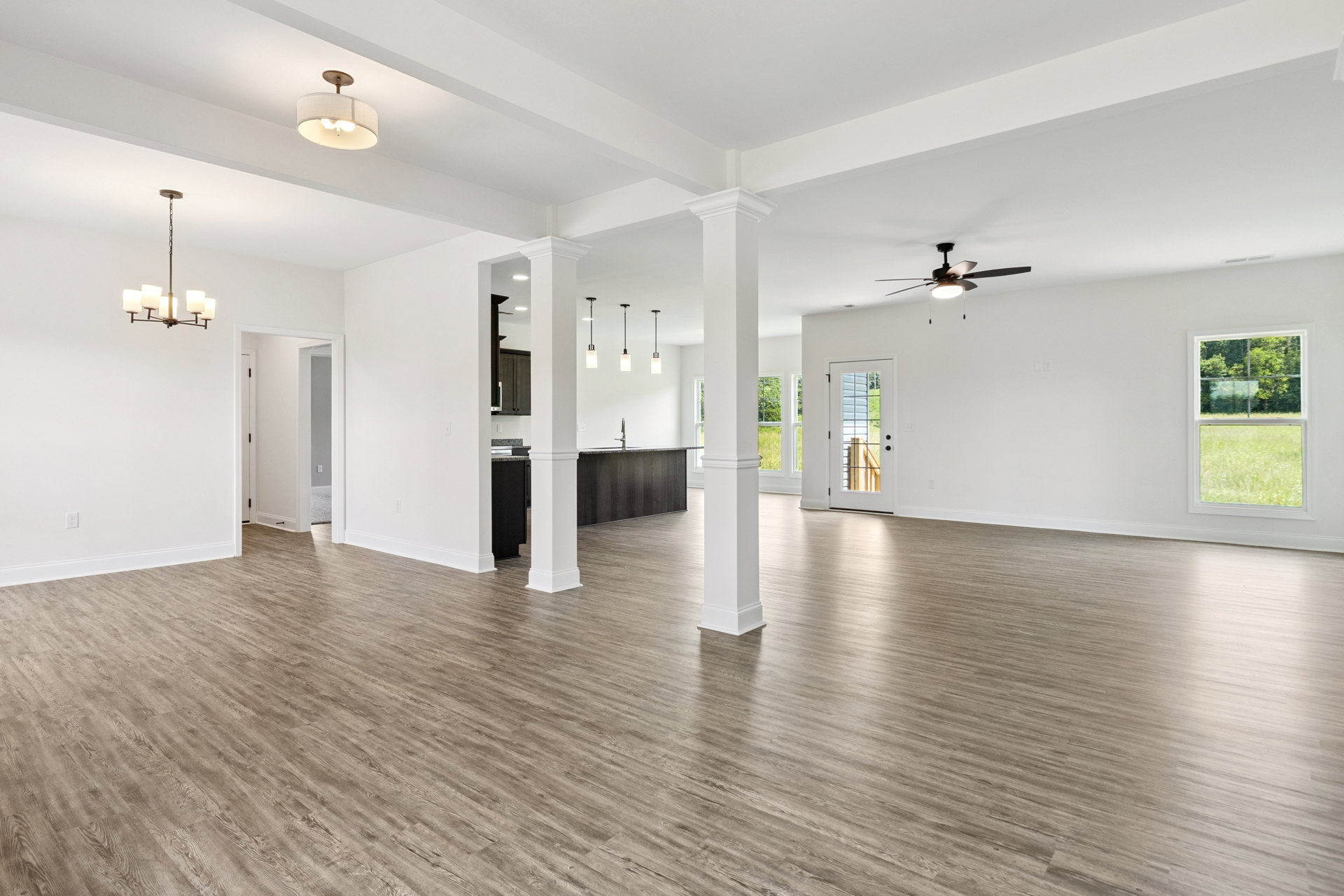 Spacious interior with white plaster walls, wood laminate flooring, white pillars, glass-paneled door, large window overlooking grassy field and trees, and a chandelier with a
