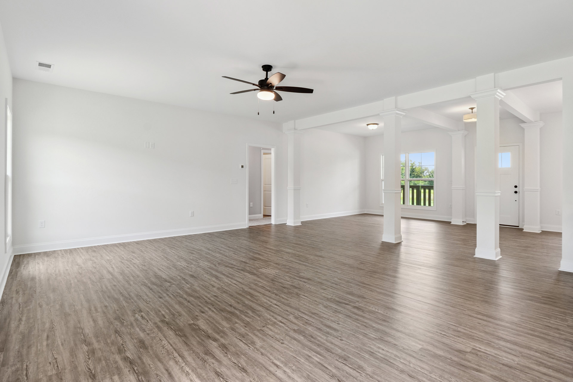 Spacious room with wood flooring, white walls, ceiling fan with light, large window showing trees outside, and white pillar near the window
