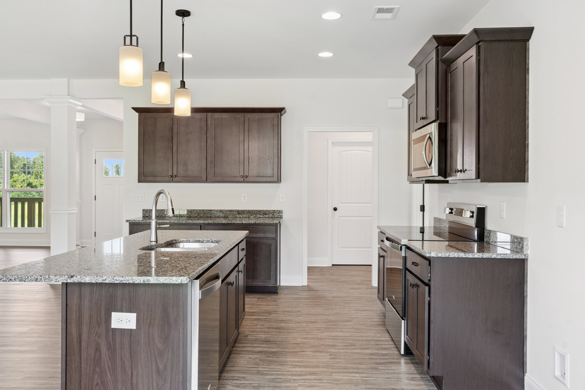 Kitchen with dark wood cabinets, granite countertops, central island with built-in sink, stainless steel appliances, pendant lighting, window overlooking trees, white electrical