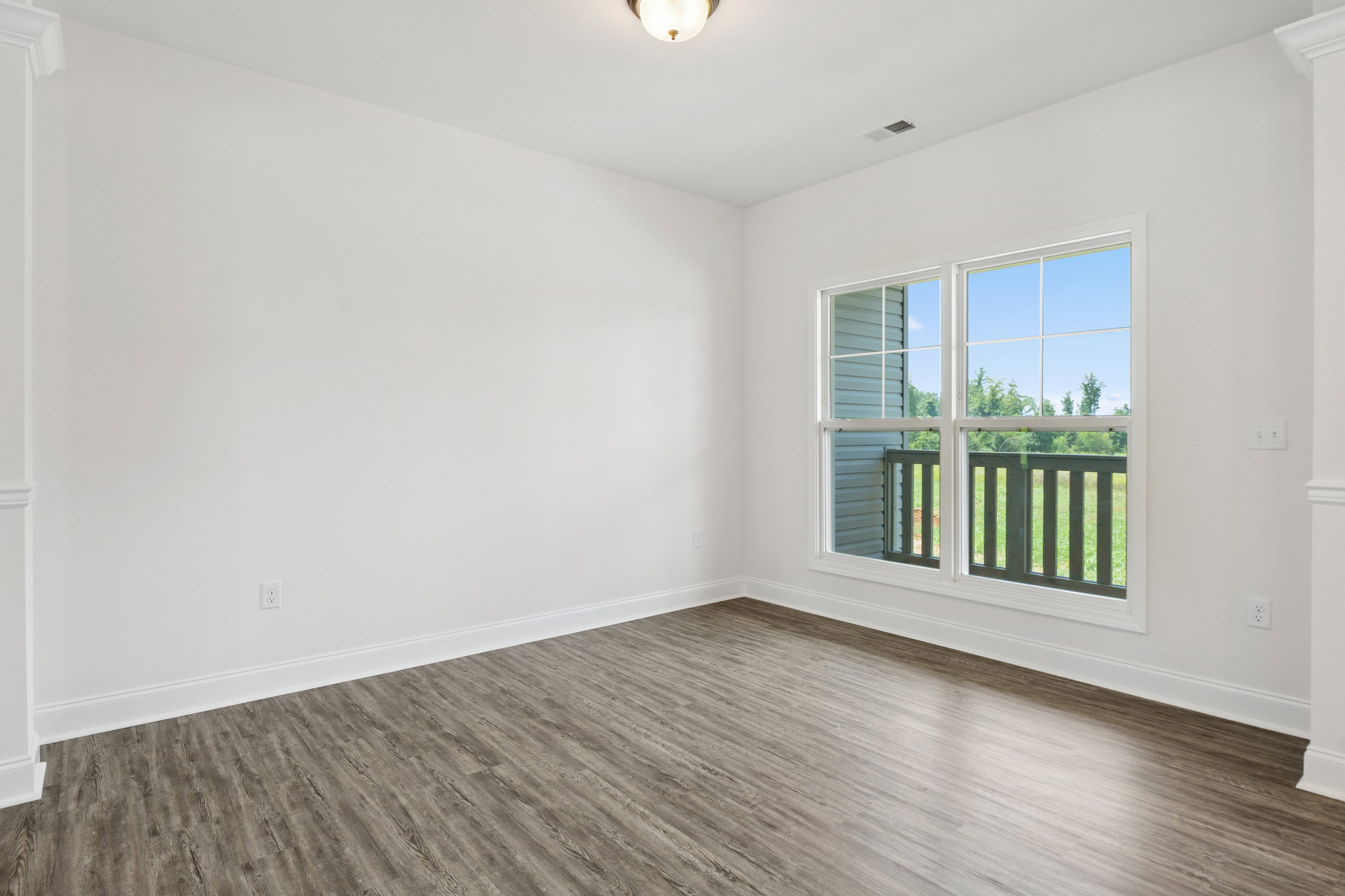 Wood flooring and white walls in a bright room with a large window and glass balcony door, overlooking a fenced grassy yard and trees.