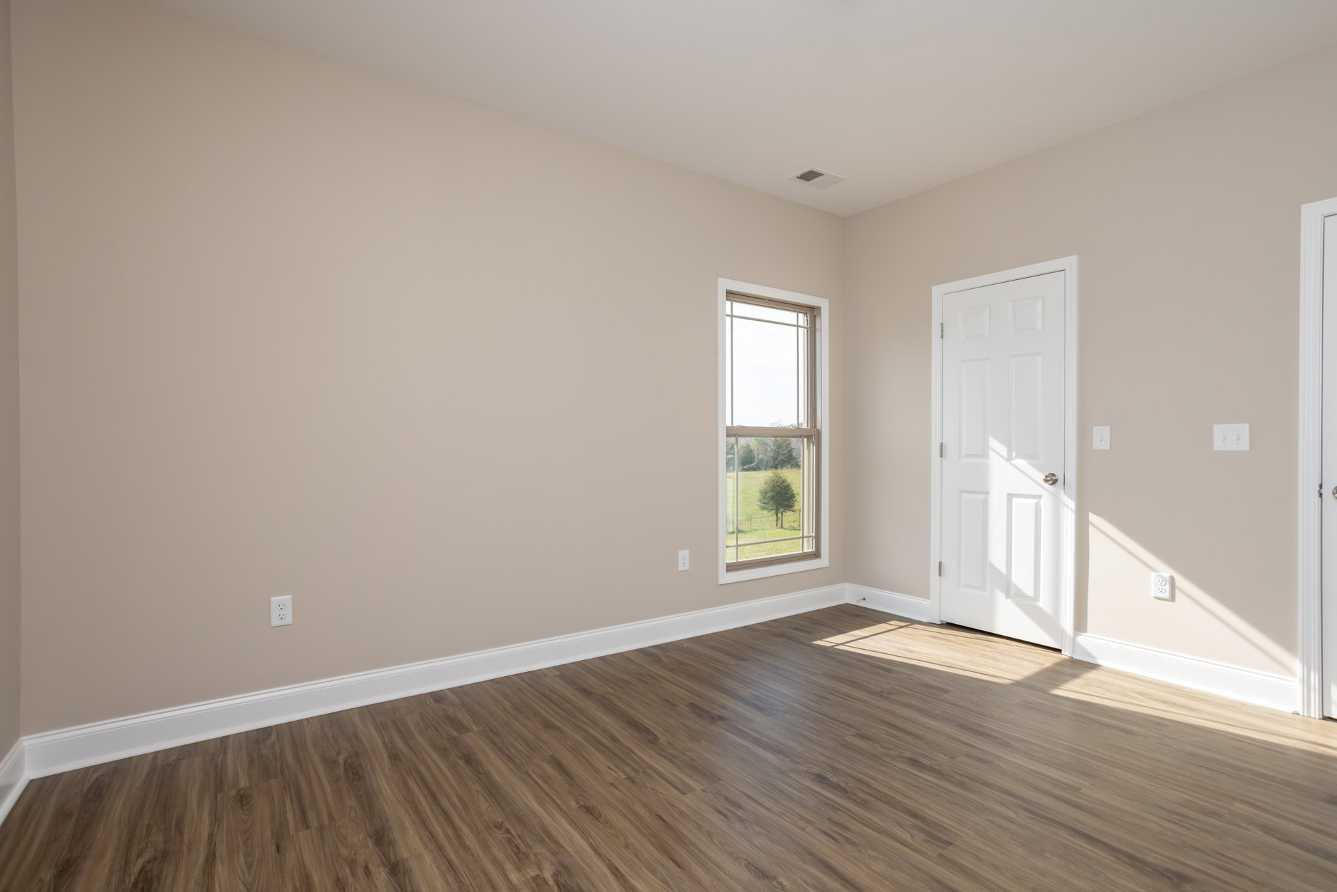 Wood flooring with white baseboard trim, white paneled door with silver knob, large window overlooking grassy field and trees