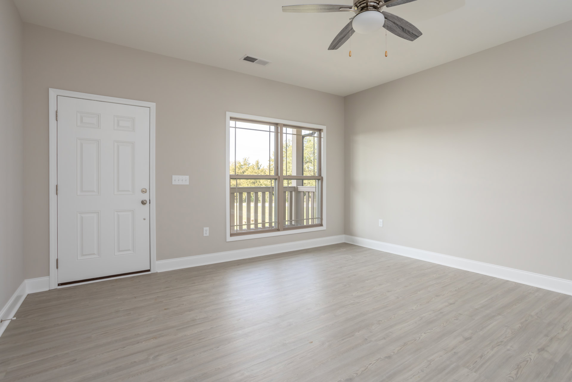 Bedroom with laminate wood flooring, white walls, ceiling fan with light fixture, white door with silver knob, window with blinds overlooking grassy field and trees.