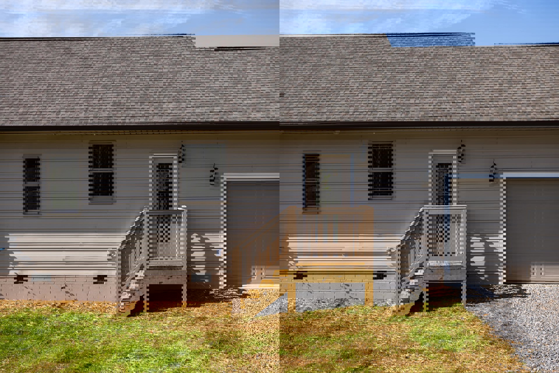 Wooden porch with railing and stairs leading to white door, white-framed window, light-colored siding, grassy yard, blue sky with scattered clouds above roof