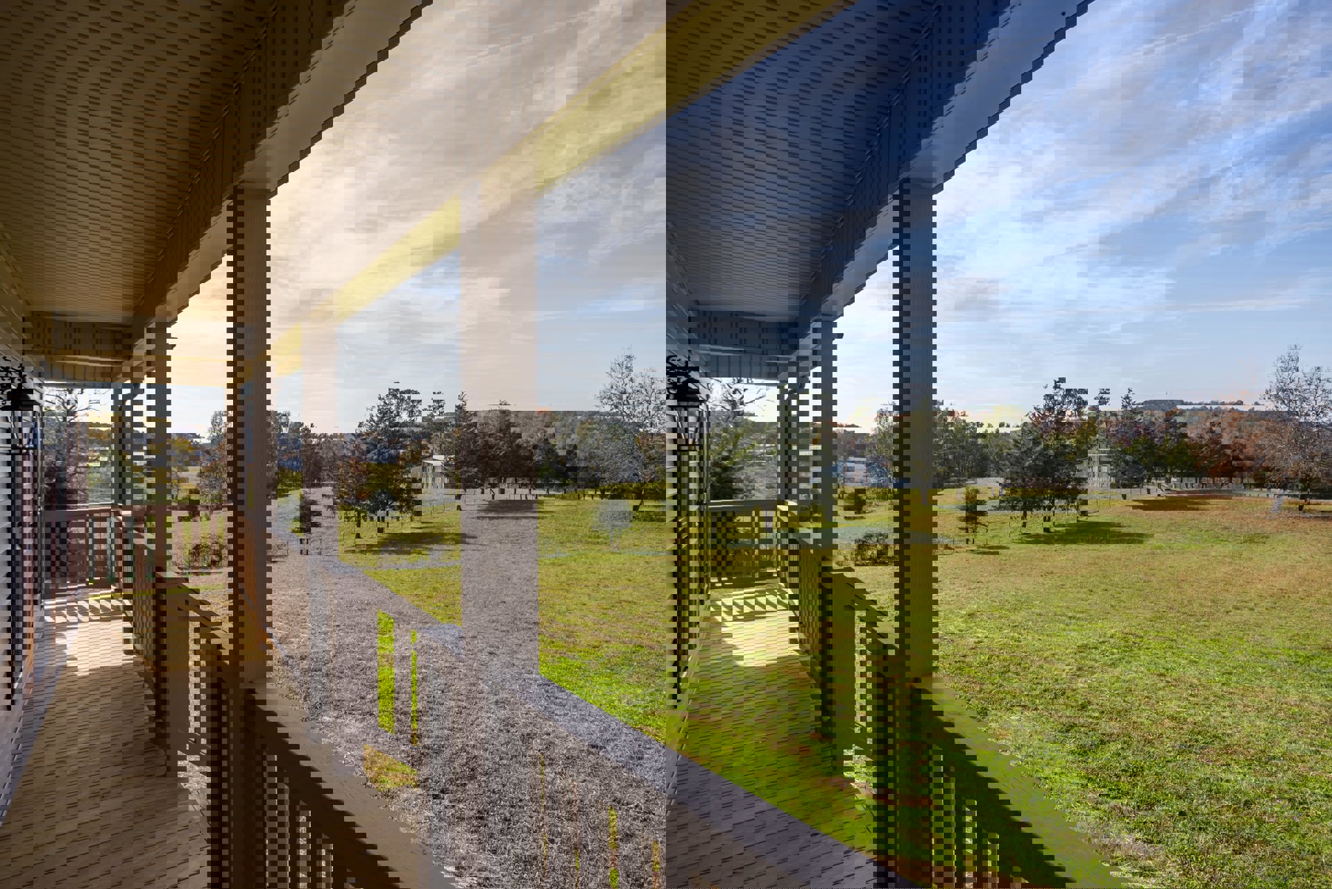 Wooden porch with white railings overlooking a grassy field dotted with trees and a distant house under a partly cloudy sky