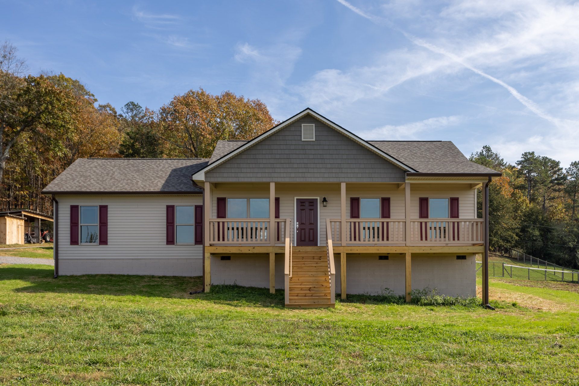 White siding house with covered porch, wooden stairs, purple shuttered windows, and green lawn bordered by trees under partly cloudy sky