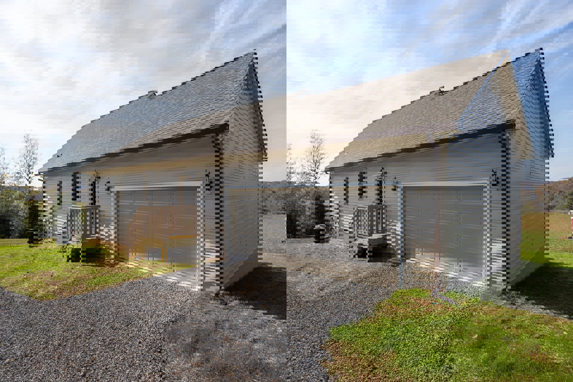 Two-story house with attached garage, gravel driveway, wooden deck with railing, grassy lawn, and surrounding trees under a partly cloudy sky