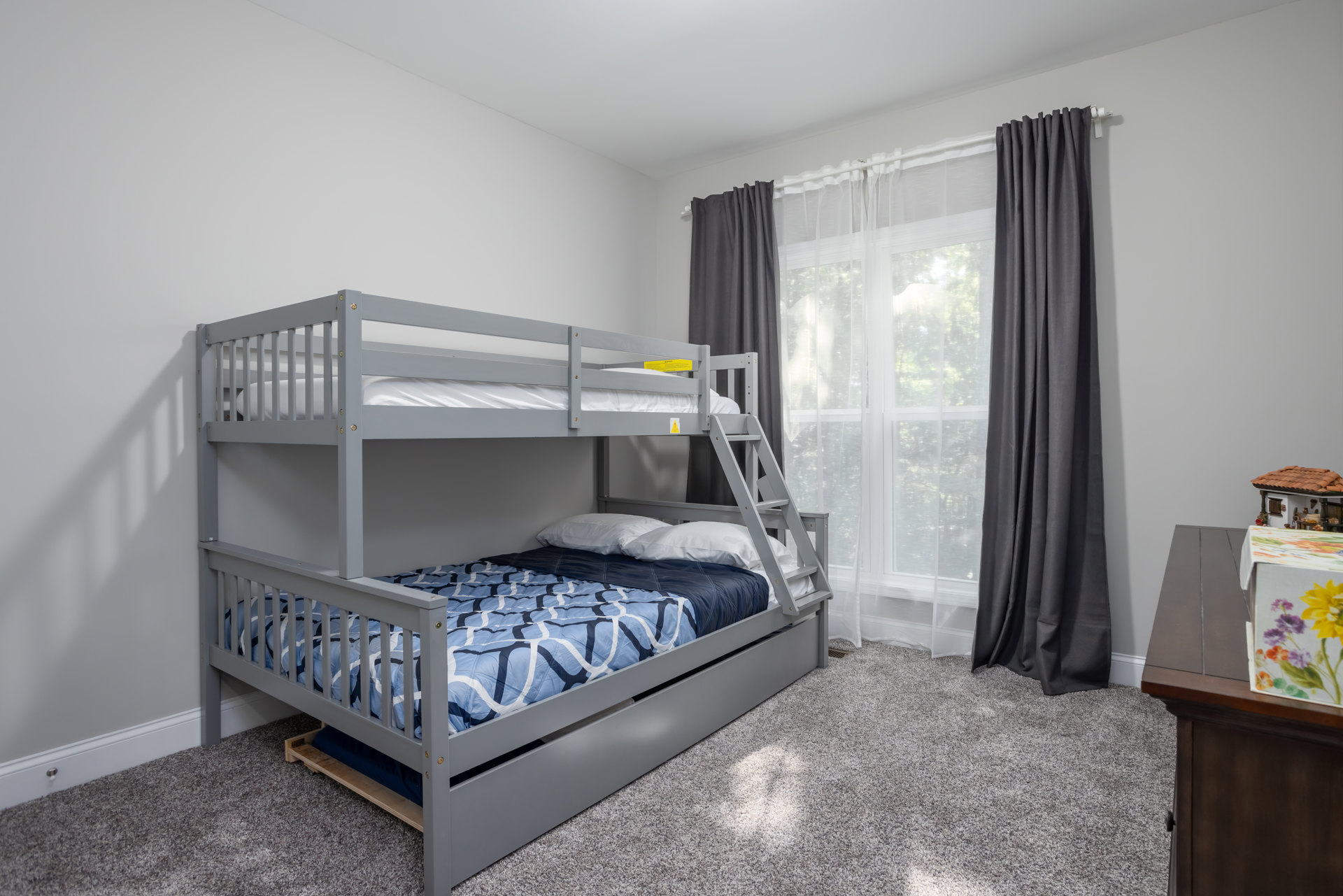 Wooden bunk bed with blue comforter and white pillows beside a window with grey curtain, light-colored walls, and hardwood floor.